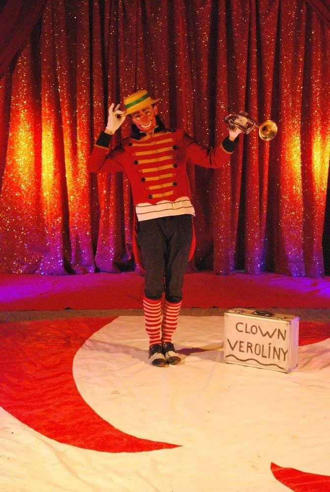 Un clown portant un costume rouge à rayures dorées et noires, un chapeau rayé vert et jaune, et des chaussettes à rayures rouges et blanches, jouant de la trompette sur scène avec un rideau de scène rouge en arrière-plan et une caisse en carton avec l'inscription 'Clown Veroliny'.