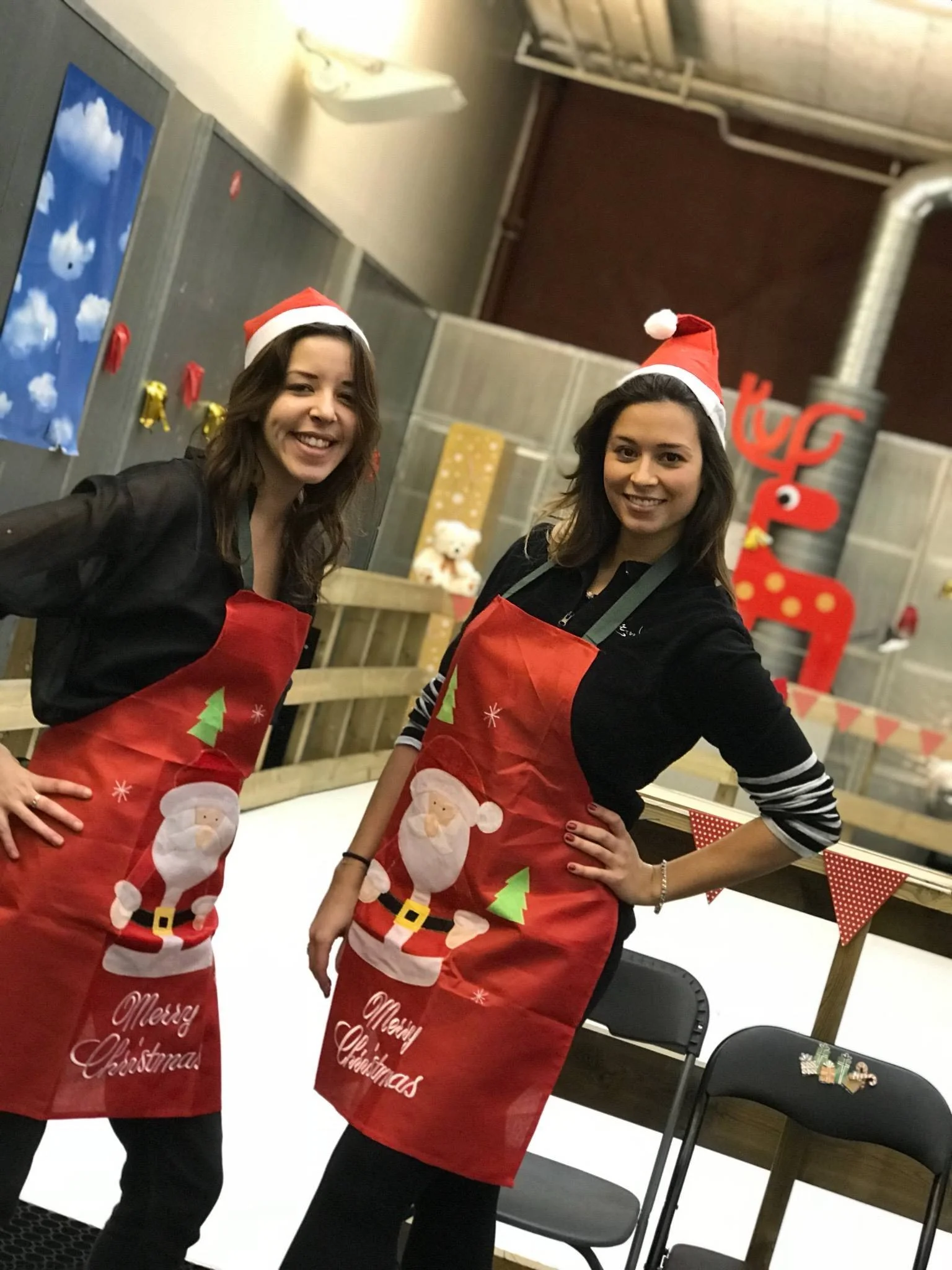 Deux jeunes femmes portant des chapeaux de Père Noël et des tabliers de Noël posent devant un décor festif de Noël.arbre de noël entreprise paris 