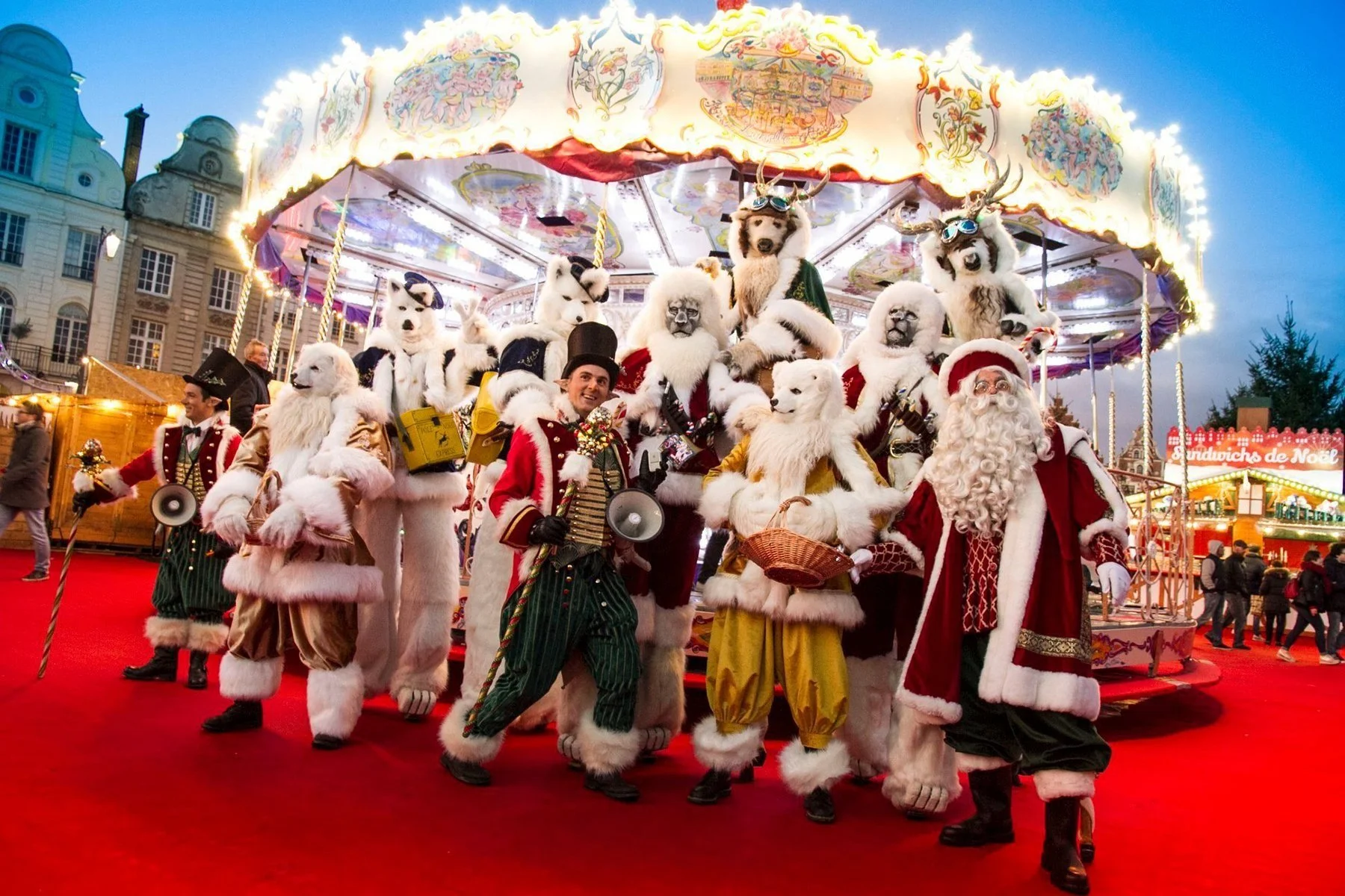 Des personnes déguisées en Père Noël et en animaux polaires, certains avec des costumes de chiens ou ours polaires, dans une fête de Noël avec un manège et des décorations lumineuses dans une ville pendant la soirée.