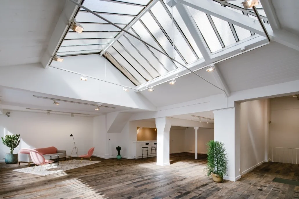 Salon intérieur spacieux avec plafond en verre, mobilier moderne avec canapé rose, fauteuils et plantes vertes, décoration minimaliste en bois