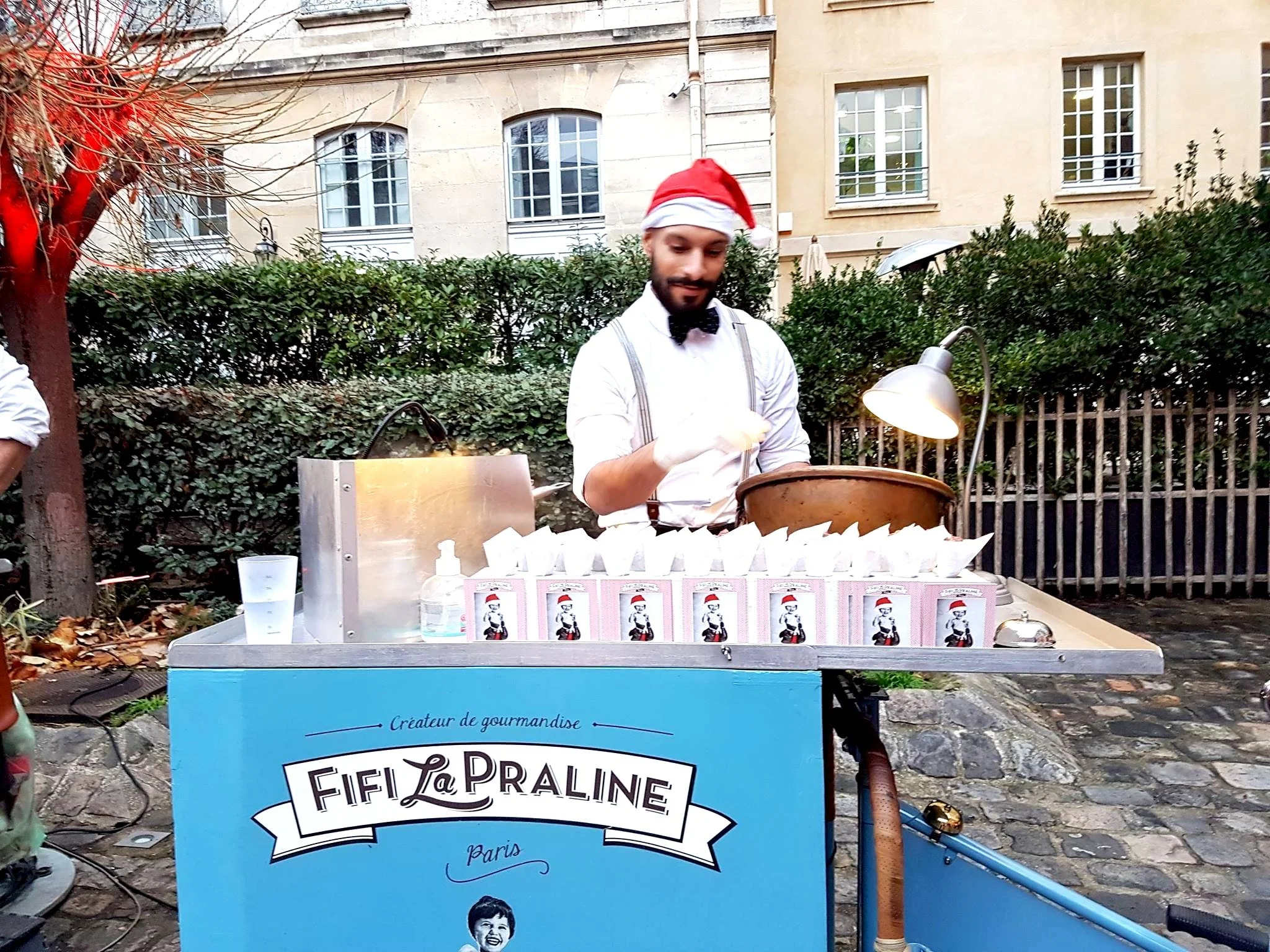 Un homme vêtu d'une chemise blanche, portant un nœud papillon noir, un bonnet de Père Noël, et des gants, prépare des bonbons ou des pâtisseries sur un stand outdoor décoré de cartons avec un logo 'Fifi La Praline' et une photo d'enfant. Le stand est éclairé par une lampe et est situé dans un espace extérieur pavé avec des buissons et des bâtiments en arrière-plan.