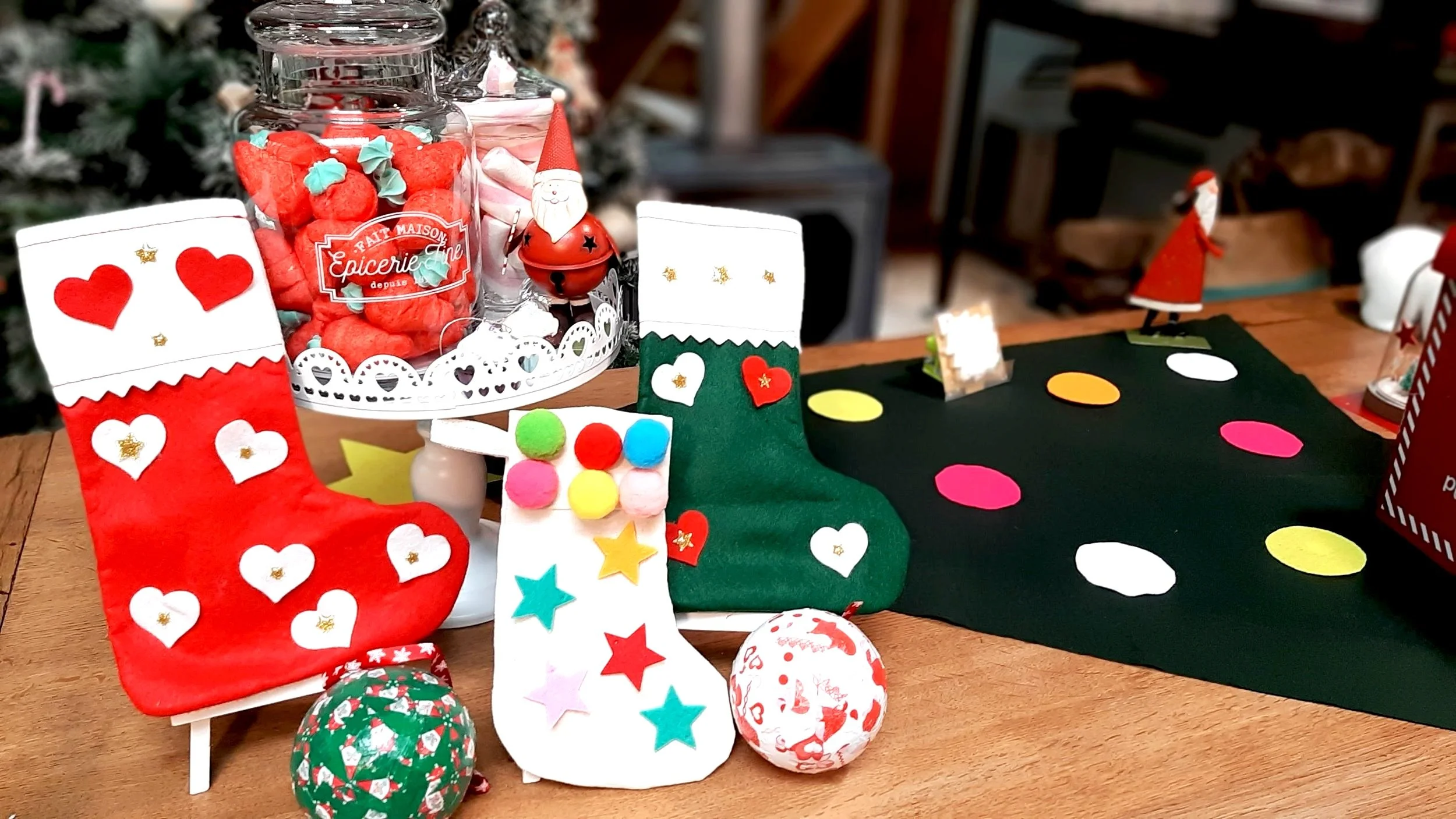 chaussette de Noël sur une table avec des chaussettes de Noël en feutrine, des figurines de Père Noël, des boules de Noël, une jarre remplie de bonbons rouges, et un tapis vert avec des points multicolores.