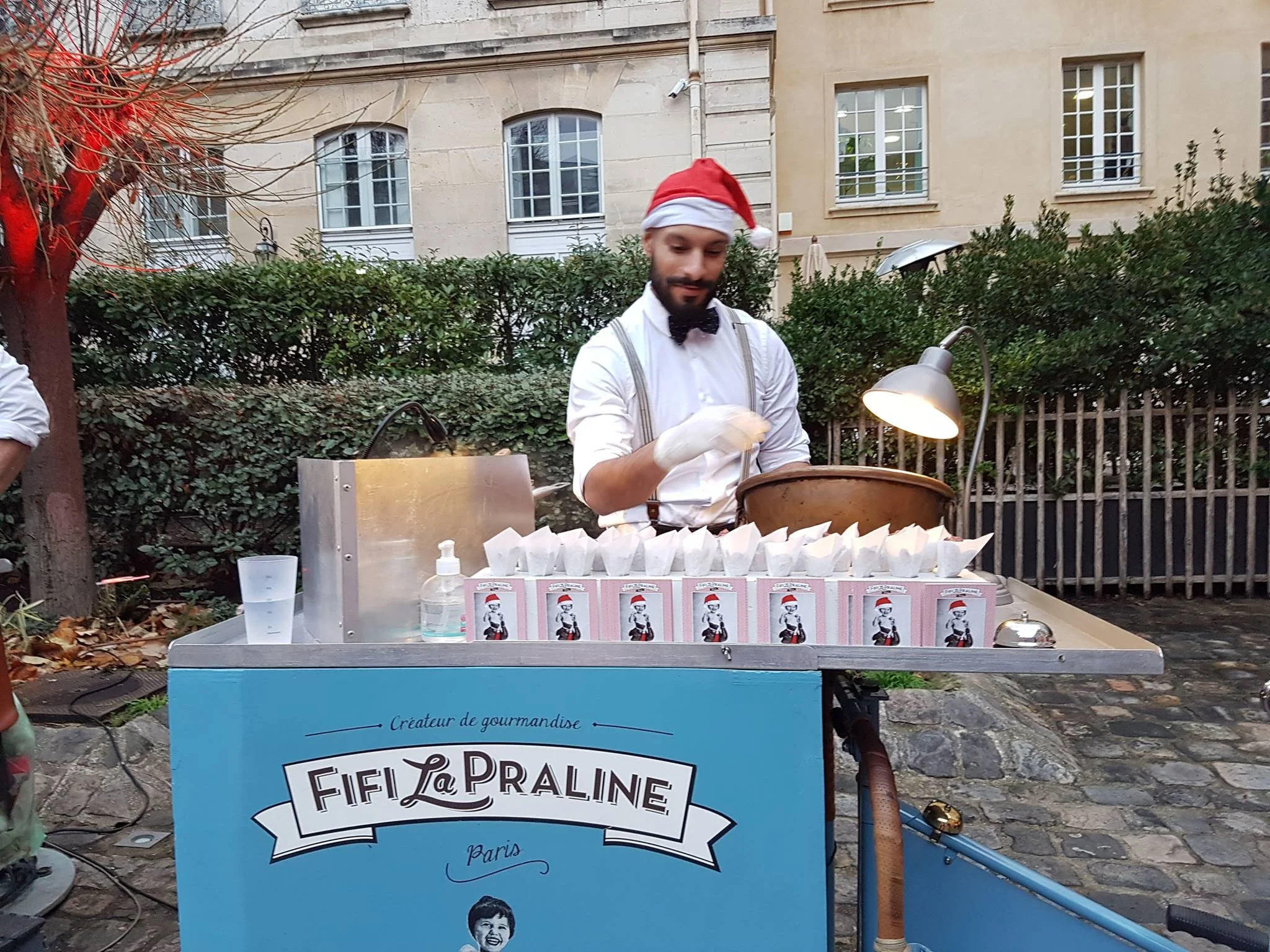 Un homme en costume avec un chapeau de Père Noël prépare des bonbons ou des friandises dans un chariot de fête foraine ou de marché de Noël à l'extérieur, avec des boissons en sachets derrière lui.