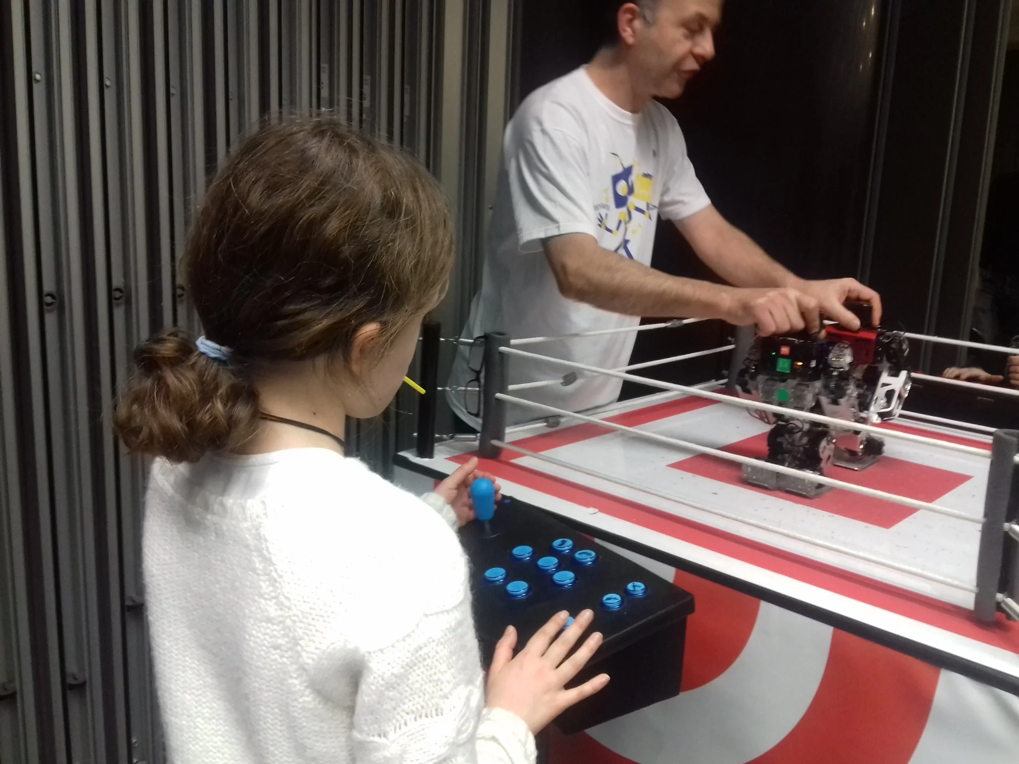 Une jeune fille utilise un joystick pour contrôler un robot humanoïde dans une arène de combat robotique, encadrée par un homme.