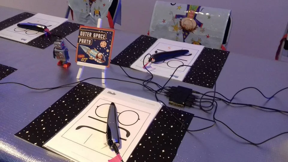 Une table de fête décorée pour une soirée d'anniversaire avec des dessins de chiffres 2 et 0, stylos, des pinces, des fils électriques, et un panneau indiquant 'Outer Space Party' avec une image d'un vaisseau spatial et une planète.