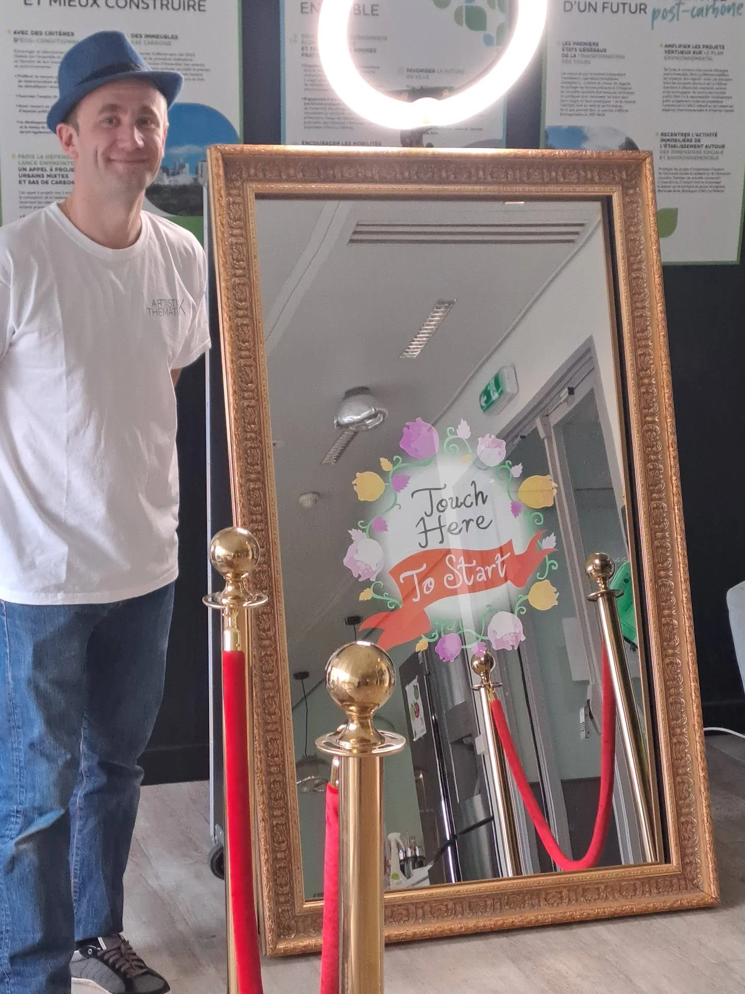 Un homme debout à côté d'un grand miroir orné d'une décoration florale, avec un panneau signifiant 'Touch Here To Start' (Touchez ici pour commencer).
