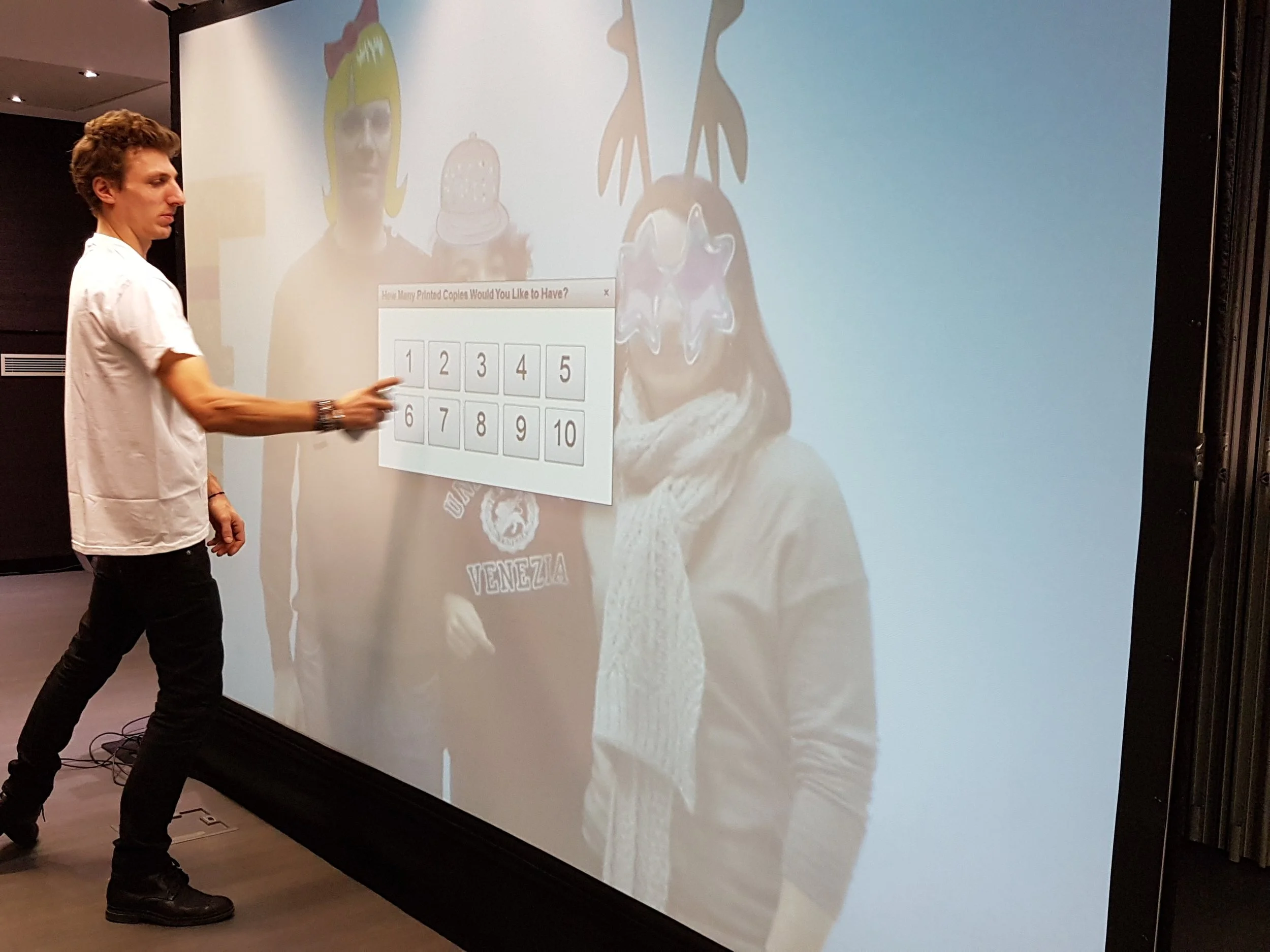Un homme pointe une zone d'interaction sur une grande toile numérique derrière lui. La toile affiche une image d'une mascotte ou d'un personnage avec des lunettes étoilées, une écharpe blanche et un t-shirt noir avec un logo. La scène semble être une présentation ou une démonstration interactive.