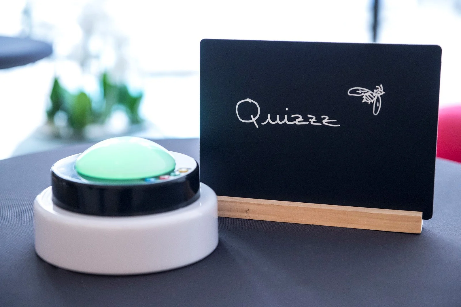 Un buzzer vert sur une base blanche, à côté d'un tableau noir avec l'inscription 'Quizzz' et un dessin d'abeille, sur une table noire.