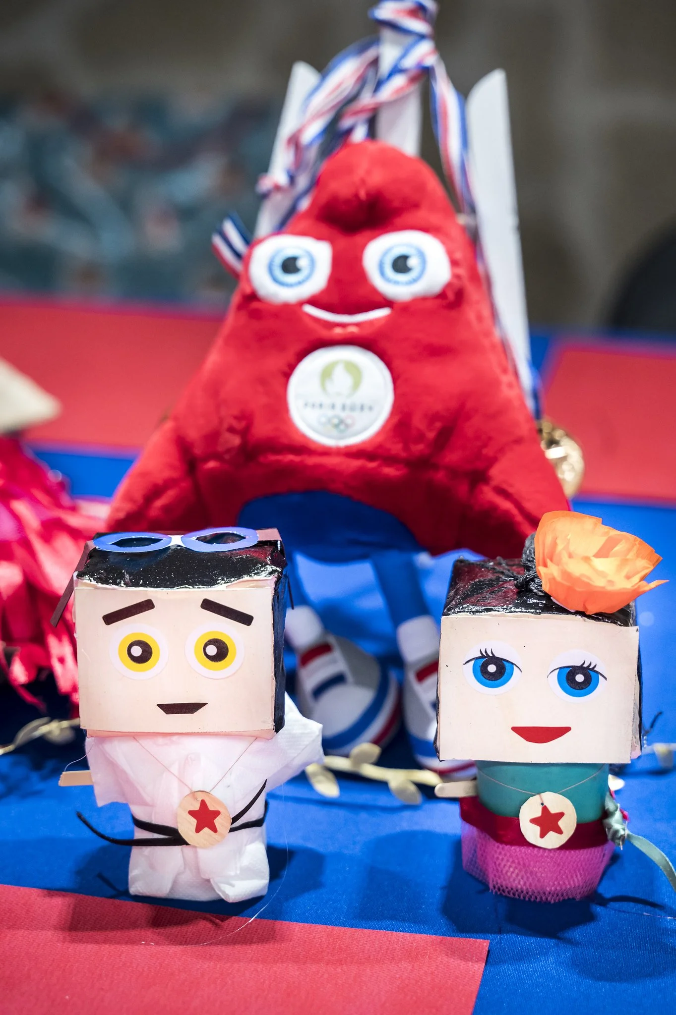 Peluches et figurines représentant des personnages avec des visages souriants et un doudou rouge, sur une table décorée de rouge et bleu.