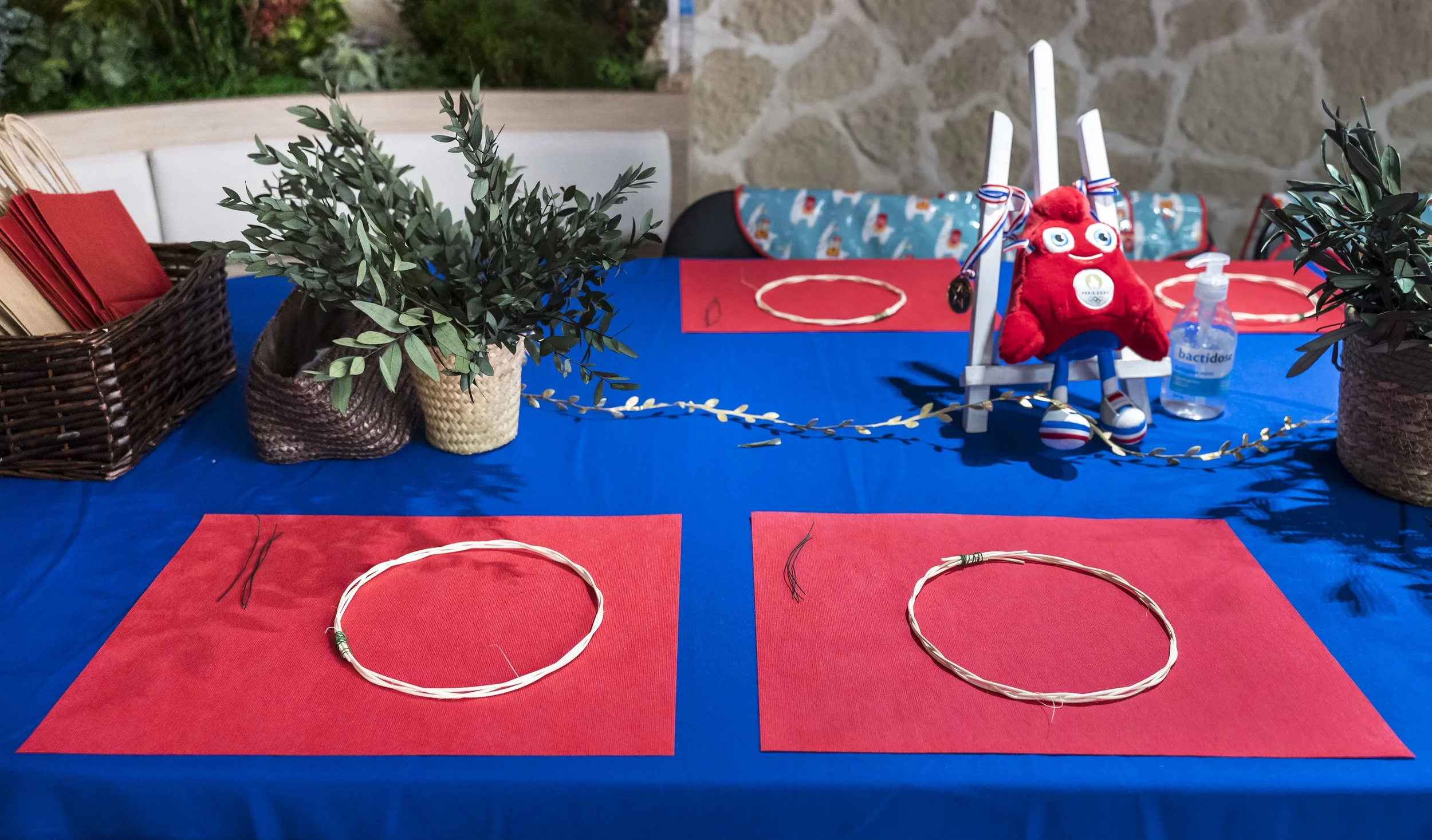 Table décorée avec un tissu bleu, avec deux places pour la création de couronnes de fleurs ou d'arts manuels, avec des plants en pot, une poupée rouge et des objets de décoration