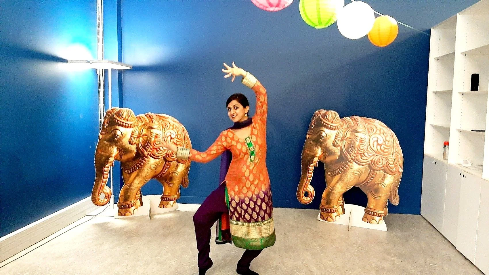 Une femme déguisée en costume traditionnel indien, posant avec deux sculptures d'éléphants dorés dans une pièce avec un mur bleu et des lampions colorés suspendus au plafond.