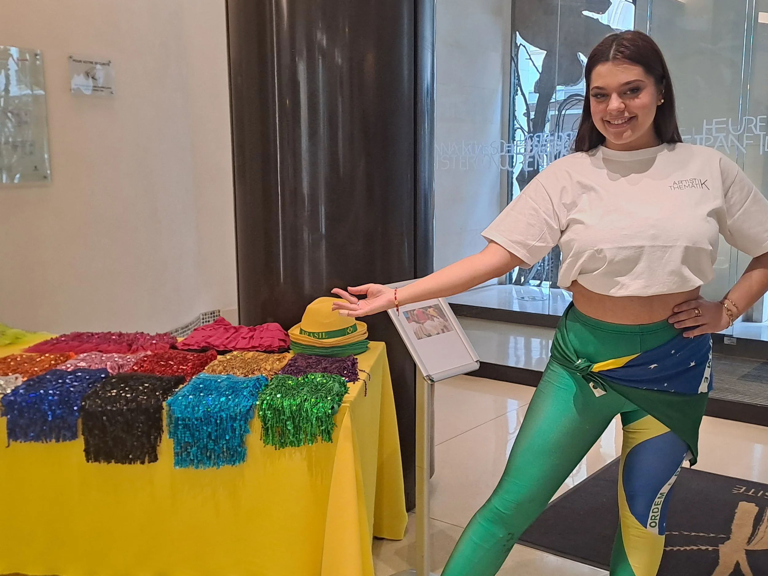 Une femme souriante portant un t-shirt blanc et un pantalon aux couleurs du drapeau brésilien se tient à côté d'un stand avec des colliers colorés et des chapeaux verts et jaunes. Elle pointe vers les accessoires sur la table.