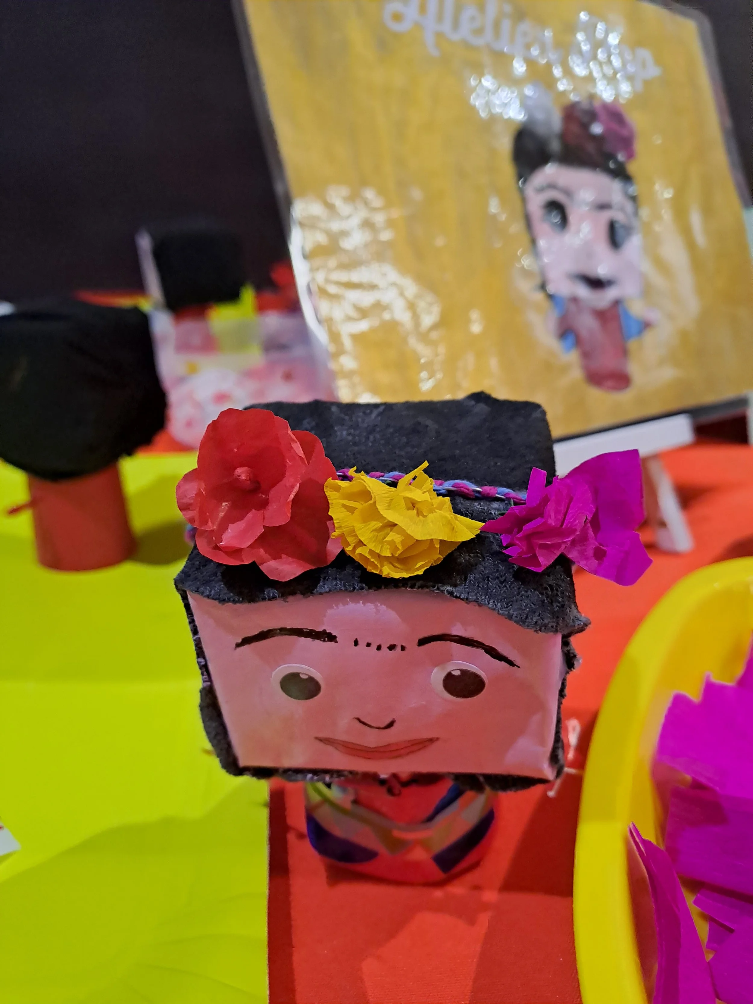 Une figurine en carton avec un visage souriant, décorée de fleurs en papier de différentes couleurs, posée sur une table colorée avec d'autres objets similaires en arrière-plan.