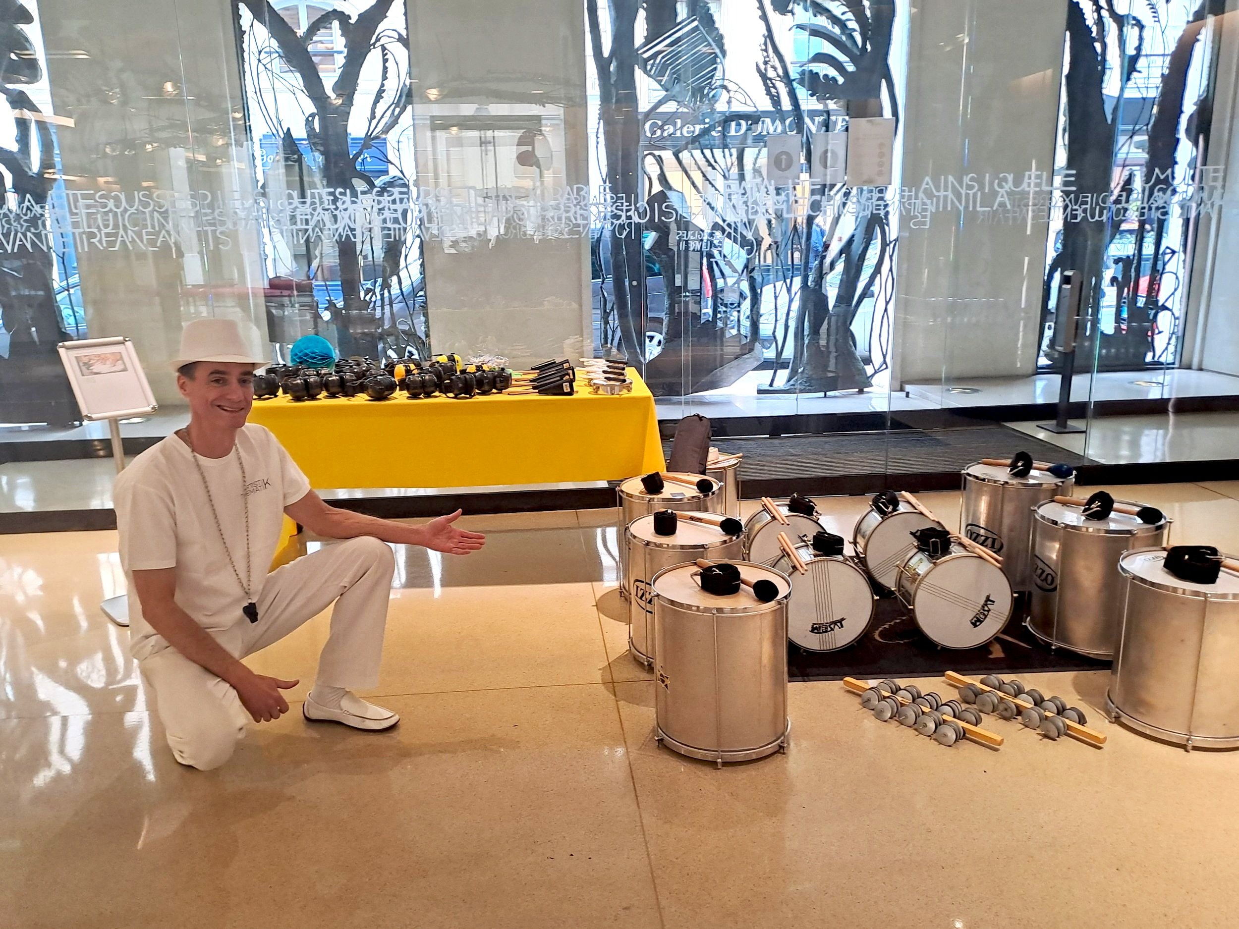 Un homme en tenue blanche, portant un chapeau blanc, souriant et gesticulant, se trouve à côté de divers instruments de musique en métal, notamment des tambours et des maracas, exposés dans un lieu intérieur avec des fenêtres qui laissent voir une rue extérieure.