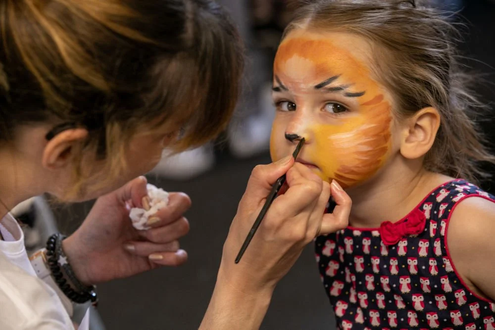 Une jeune fille se fait maquiller un visage de tigre par une artiste, lors d'une activité artistique.