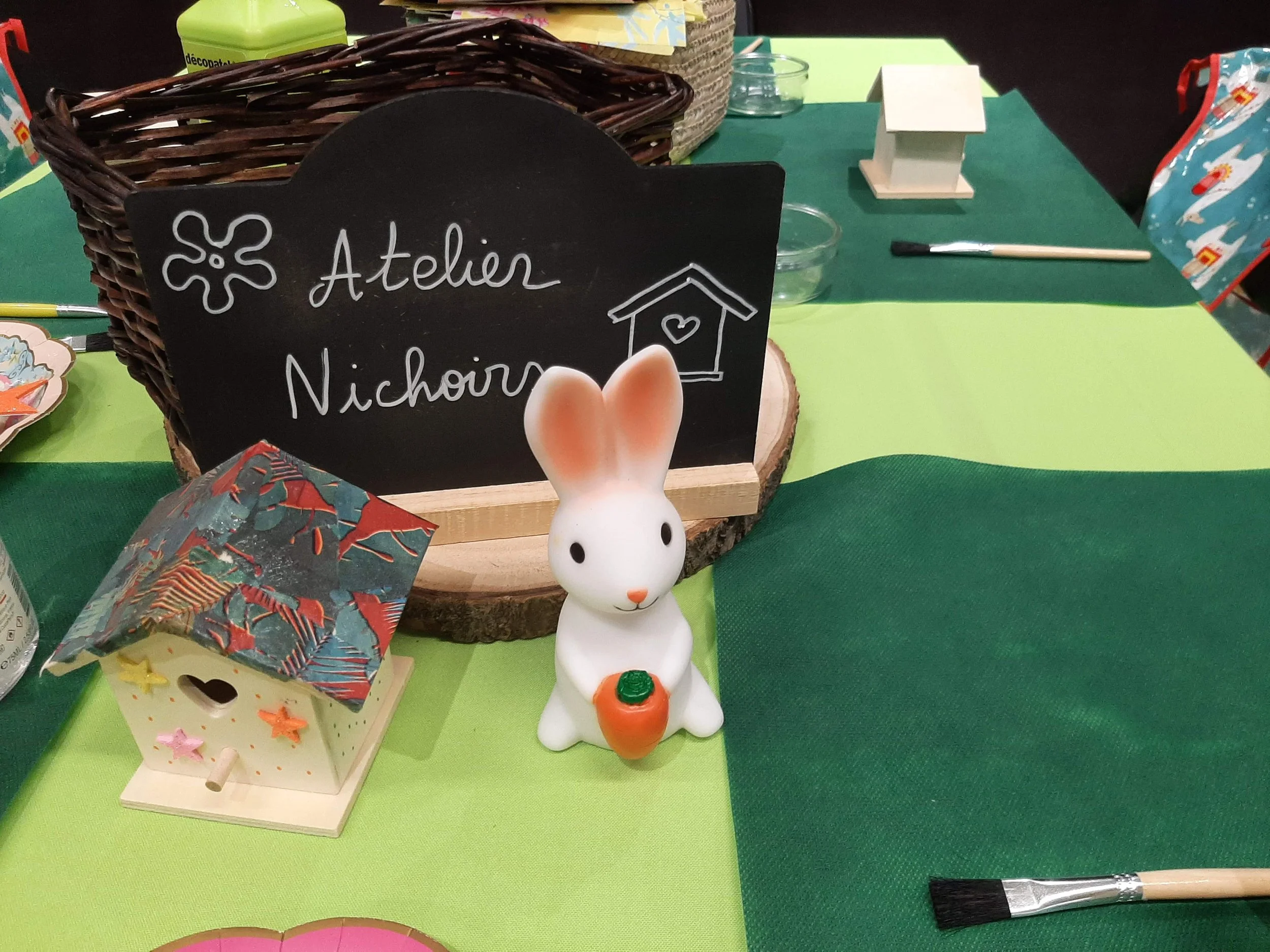 Un panneau noir avec l'inscription 'Atelier Nichoir' écrit en blanc, un petit éléphant en céramique tenant une carotte, une maison en bois décorée de petites étoiles, et un décor de table vert et gris à l'arrière.