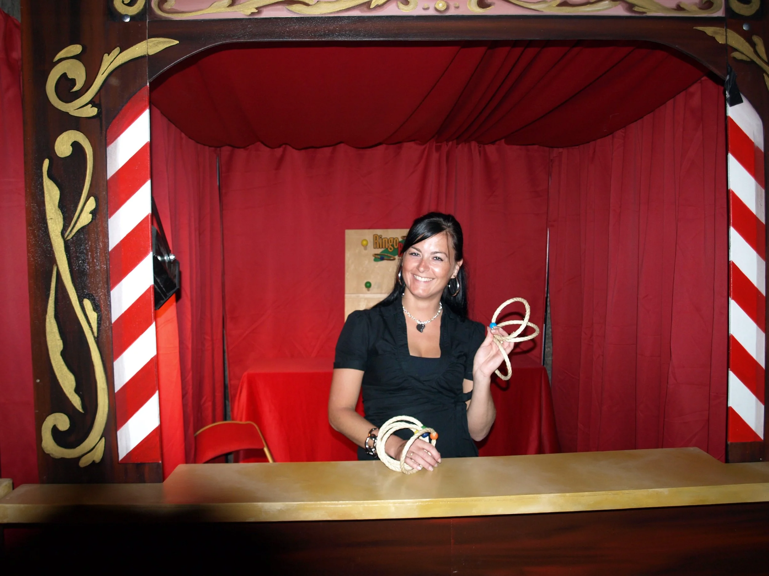 Une femme souriante derrière un comptoir rouge dans une cabine de jeu de candy game, tenant une corde en main.