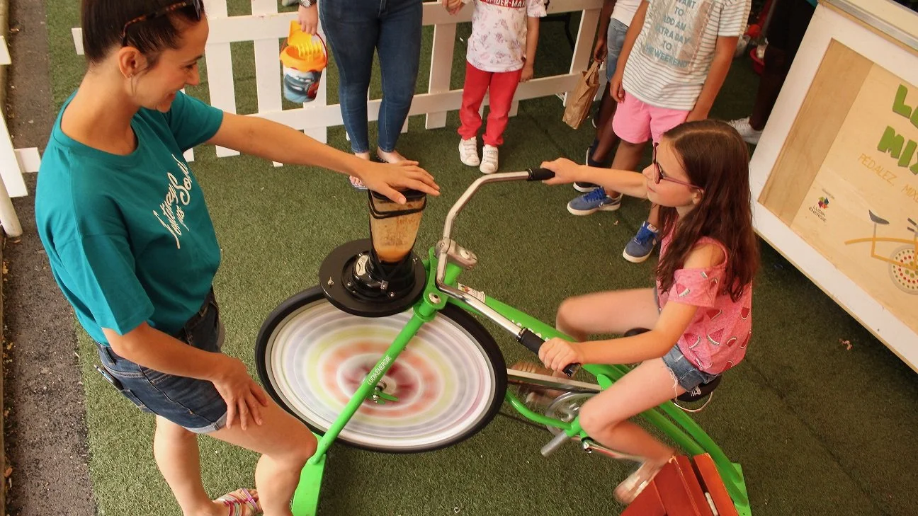Une jeune fille en train de faire du vélo stationnaire, participant à une activité de smoothie, sous la supervision d'une femme, avec plusieurs enfants observant, dans un espace extérieur
