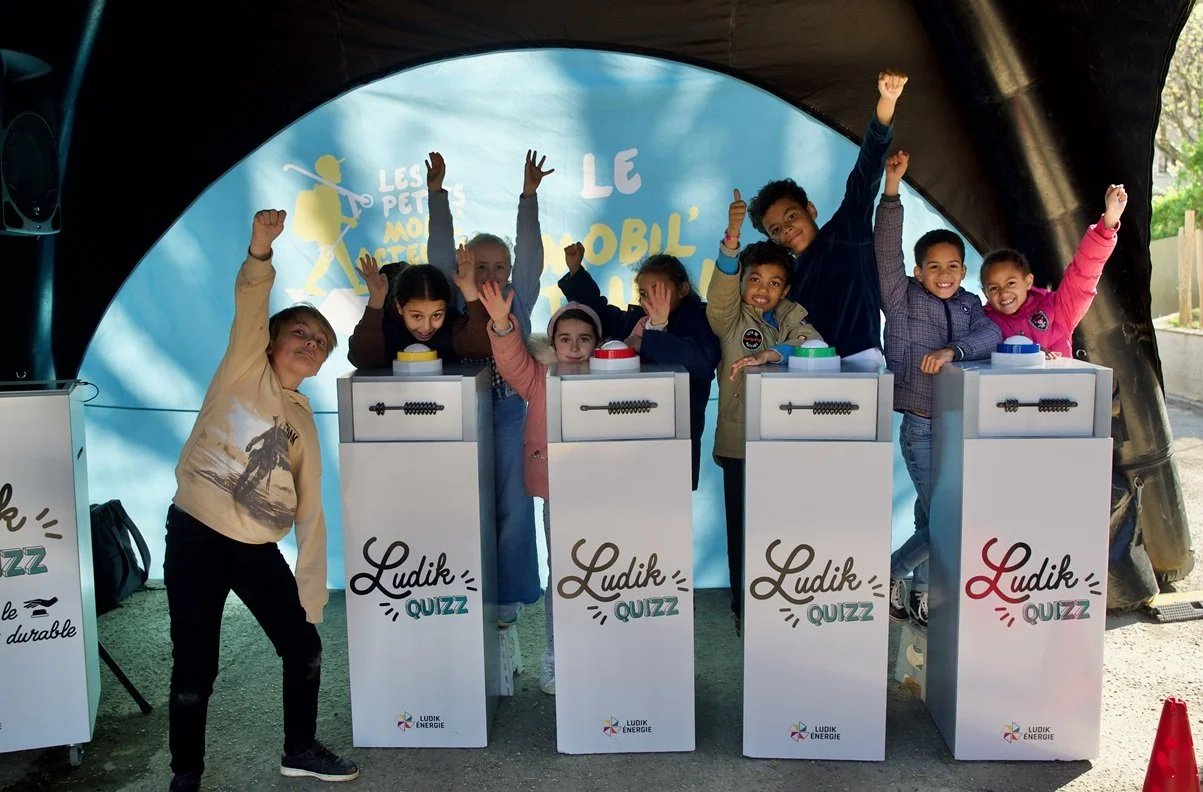 Groupe d'enfants souriants et levant la main lors d'une activité de quiz, sous une tente, avec un arrière-plan avec le texte 'LudiK QUIZZ'.
