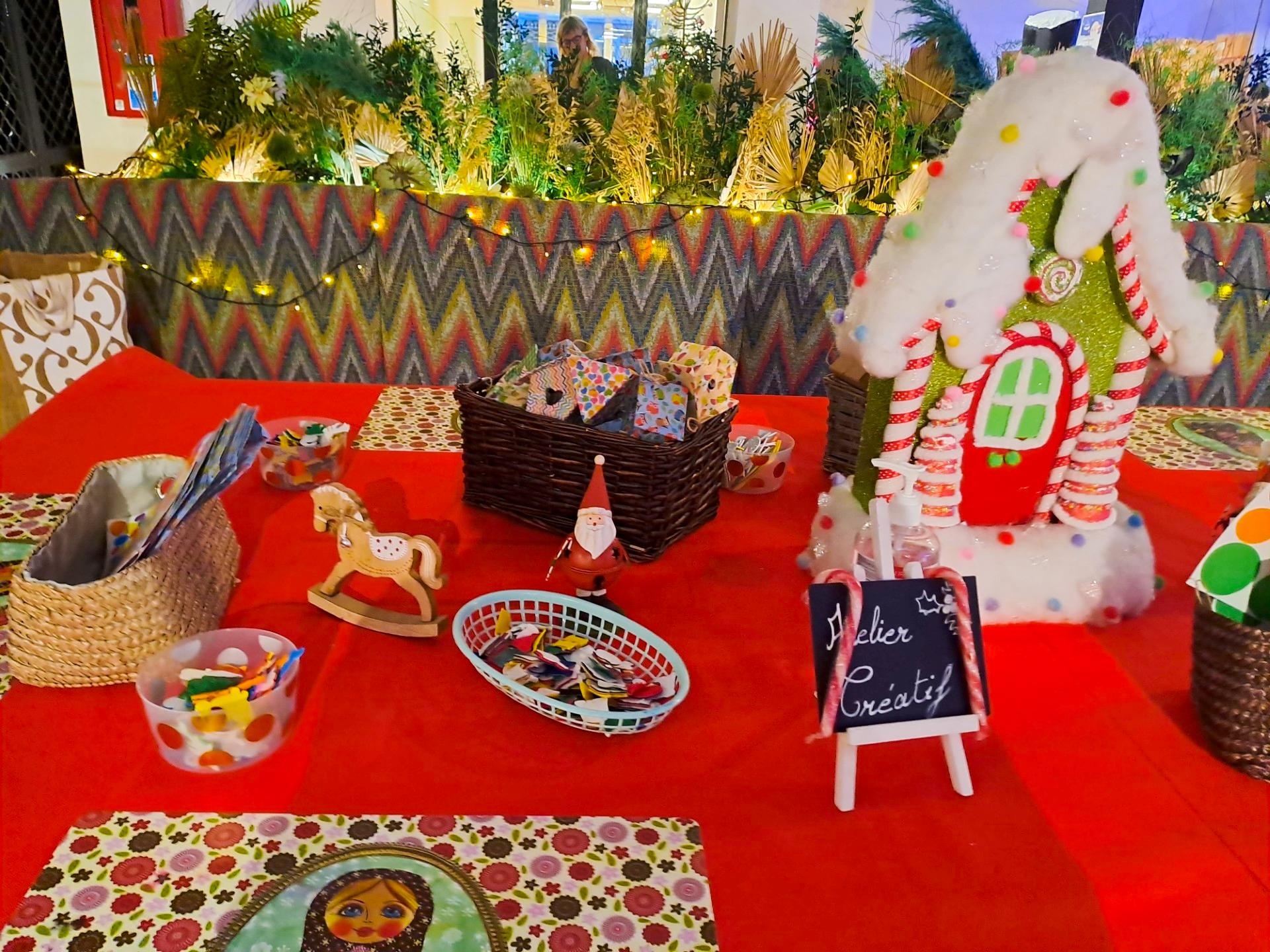 Table décorée pour Noël avec un centre de table en forme de maison en pâte à sucre avec des rubans candy, un panneau 'Atelier Créatif', plusieurs petits paniers avec des fournitures, et une décoration de style Noël en arrière-plan.