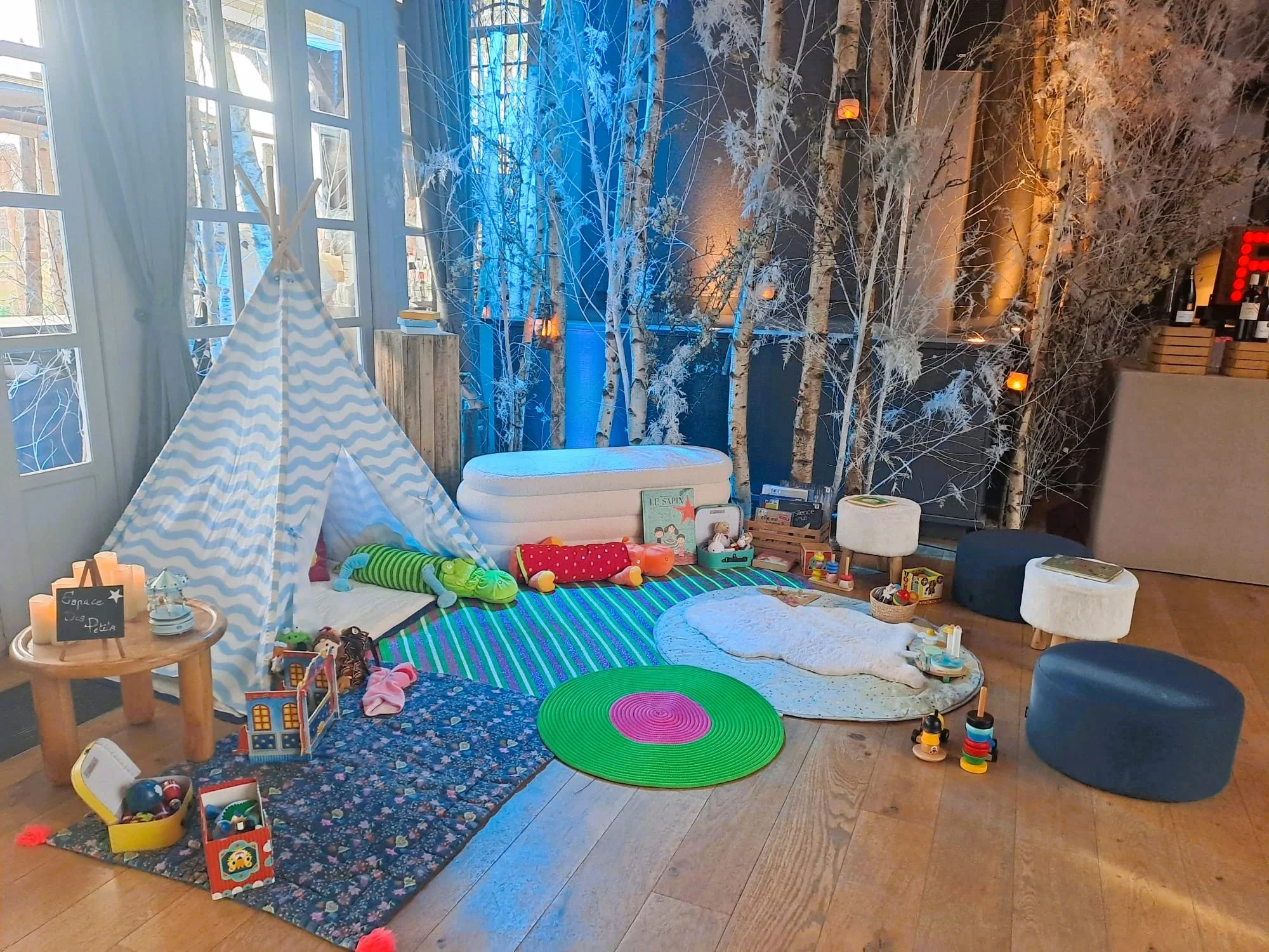 Espace de jeux pour enfants avec tente, jouets, tapis colorés, et décor de branches d'arbres blanches avec lumière bleue et orange dans une pièce lumineuse.espace petite enfance family day kidsday 