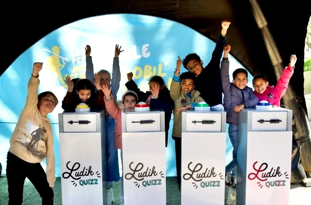 Groupe d'enfants souriants et levant les bras, jouant à un quiz Ludik Quiz sous une tente.