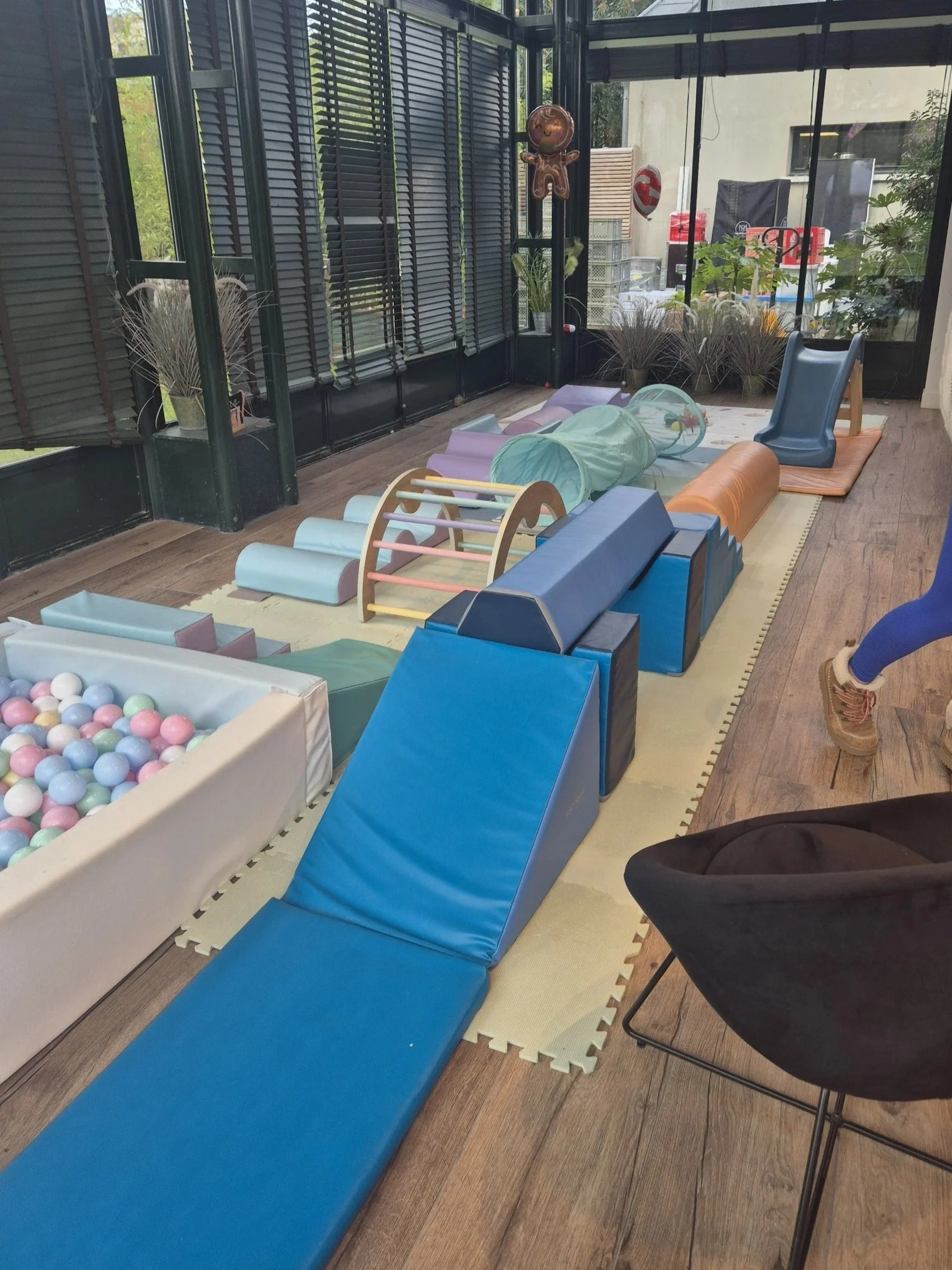 Espace de jeux pour enfants avec toboggans, tunnels, balles colorées, et tapis de jeu, dans une salle avec grandes fenêtres et décorations
