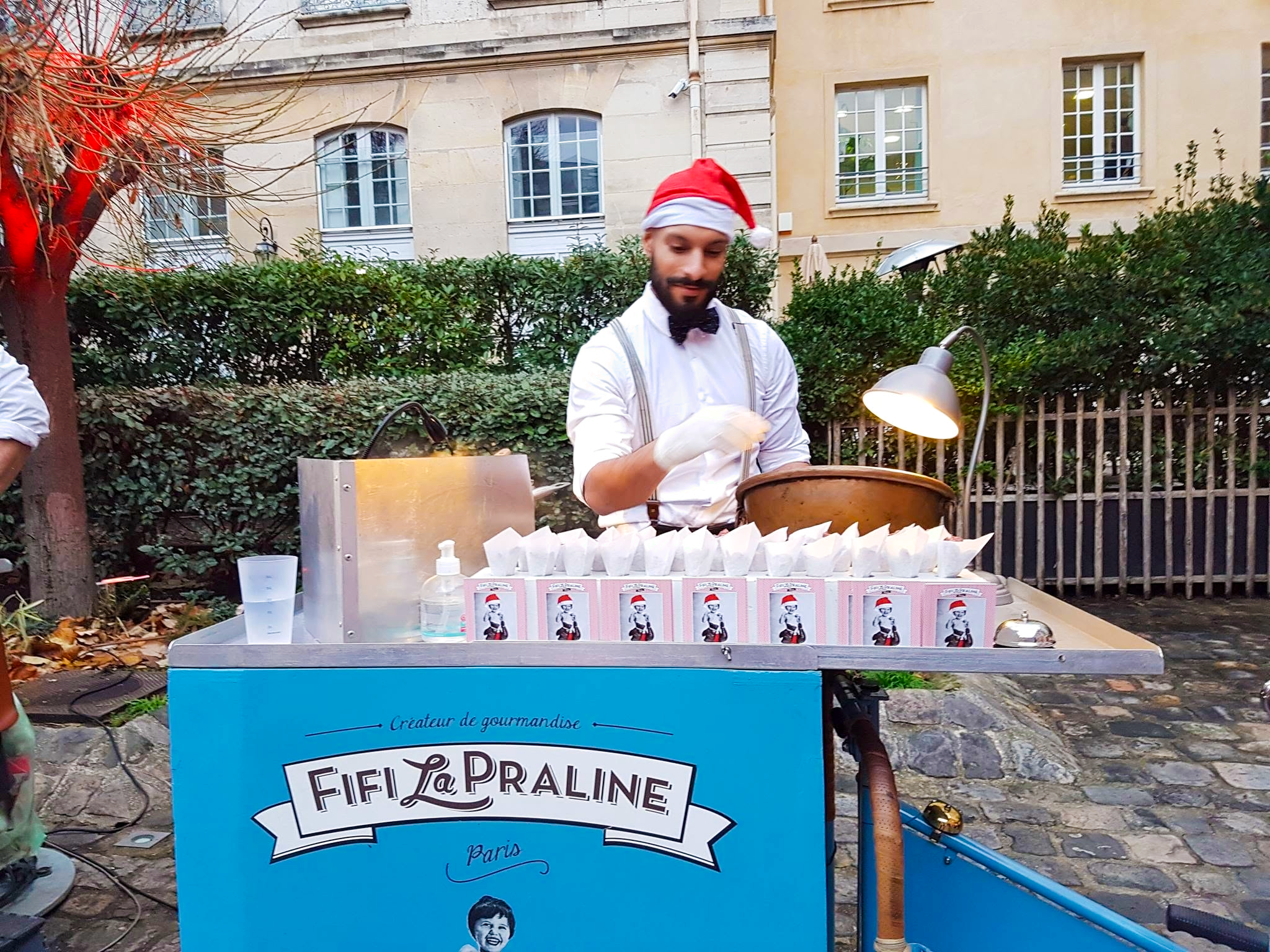 Un homme portant un chapeau de Père Noël, une chemise blanche et une cravate papillon prépare des friandises dans un stand en extérieur. Le stand est décoré avec une enseigne bleue indiquant 'Fée La praline Paris'. Il y a des petits sacs en papier devant le stand.