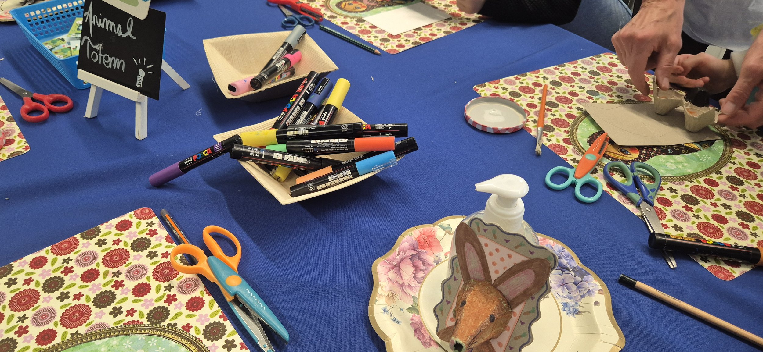 Table de fête avec des fournitures d'art et décorations colorées. On voit des marqueurs, des ciseaux, des plates à cupcakes, et des papiers décoratifs avec un thème floral. Une personne est en train de découper quelque chose.