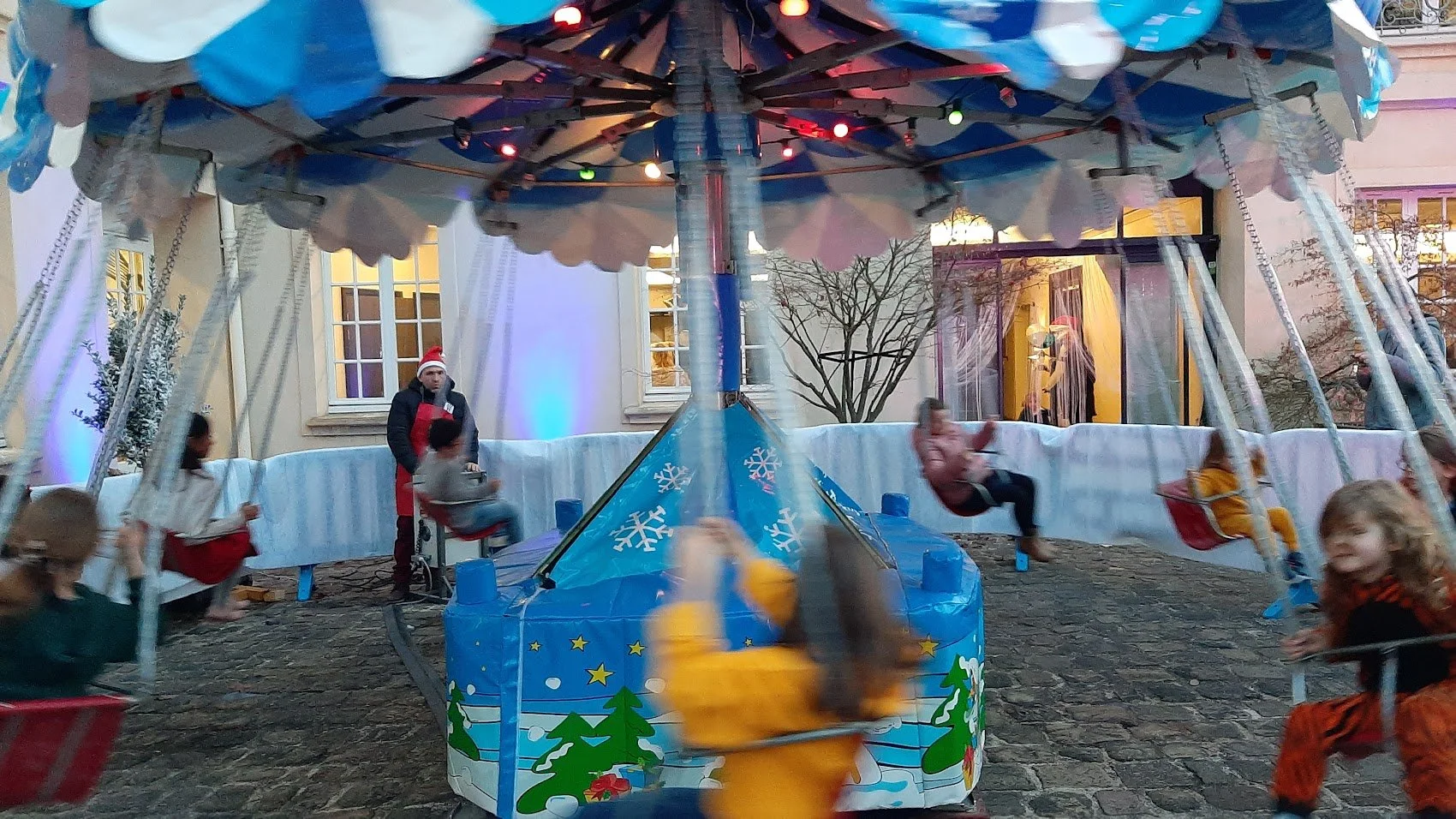 Roue de la fête foraine avec des enfants qui tournent sur les sièges et un homme avec un bonnet de Noël à côté, décorations de Noël, bâtiment en arrière-plan.