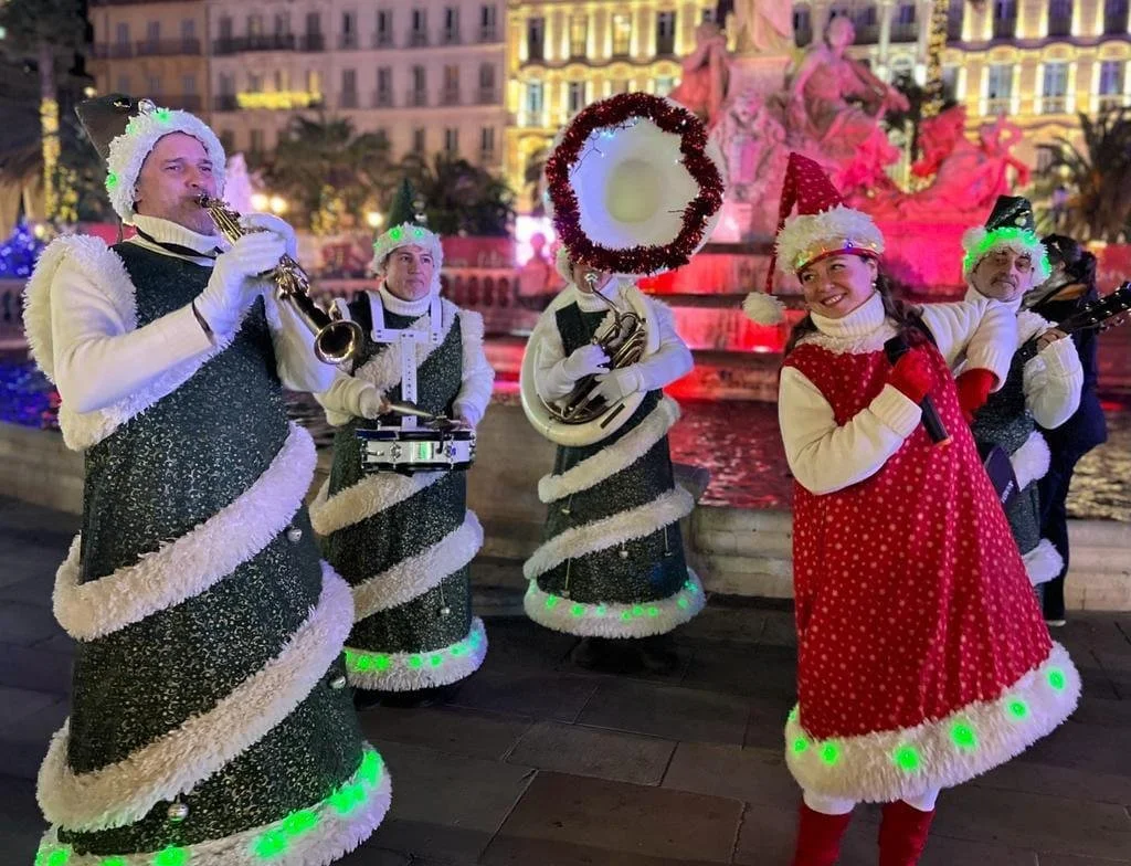 Groupe de musiciens déguisés en costumes de Noël avec des chapeaux de Père Noël, jouant de différents instruments devant une fontaine décorée, avec un bâtiment illuminé en arrière-plan