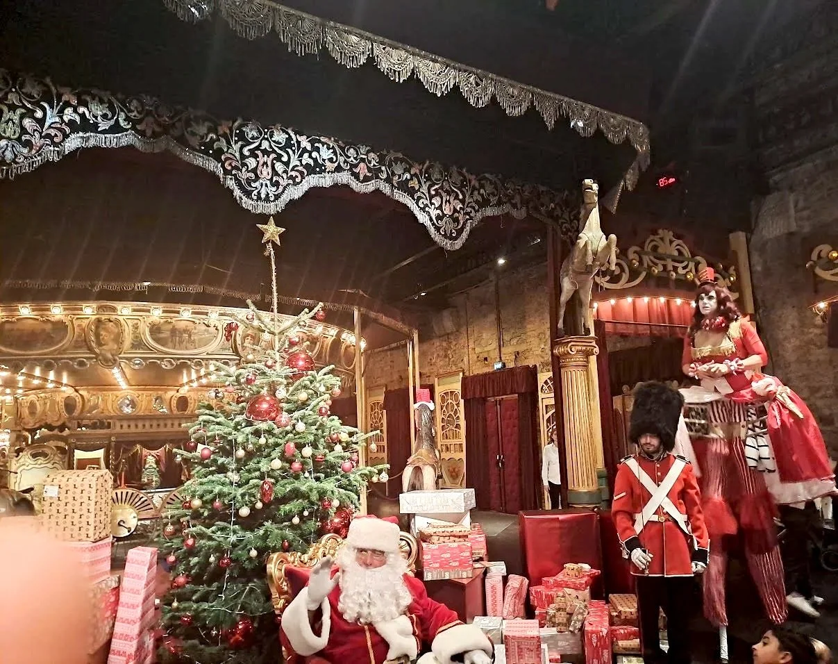 Décor de Noël avec un sapin orné de boules rouges et or, un Père Noël assis à côté, présents autour, décorations festives, figurines de soldats et un cheval en bois dans un lieu festif.
