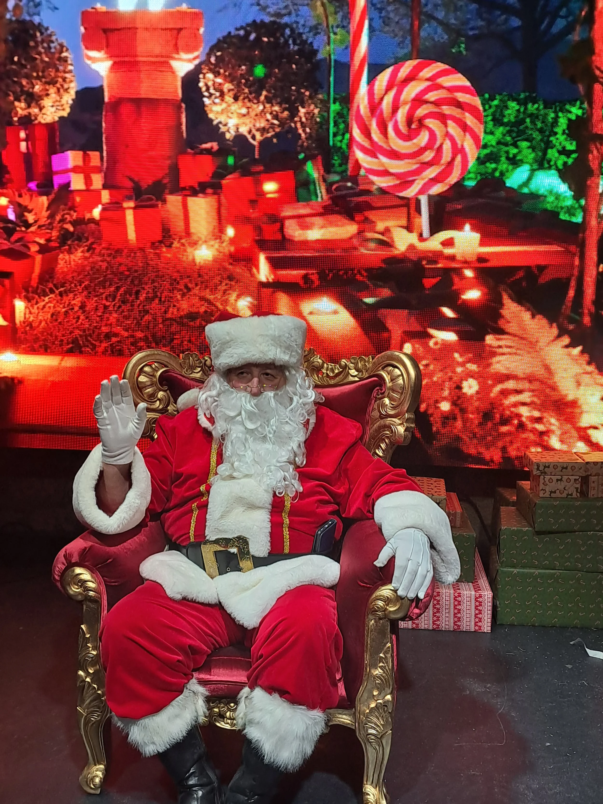 Père Noël assis sur un fauteuil doré, faisant un signe de la main, avec un décor de Noël en arrière-plan incluant des cadeaux, un bonhomme de pain d'épice géant, des sapins et des guirlandes.