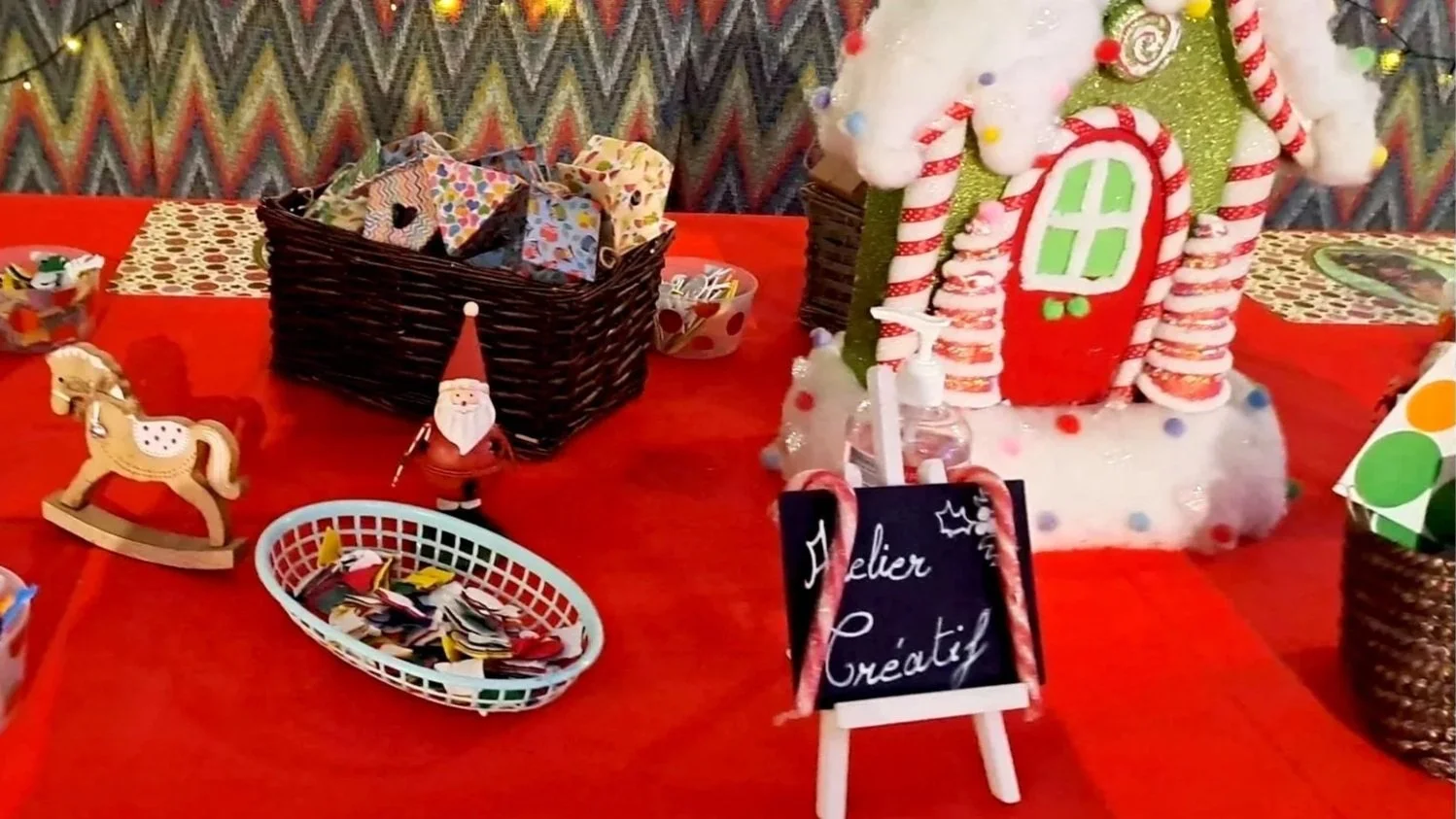 Décoration de Noël avec une petite maison en feutrine, un sapin de Noël miniature, et des paniers de cadeaux et décorations déposés sur une table rouge.