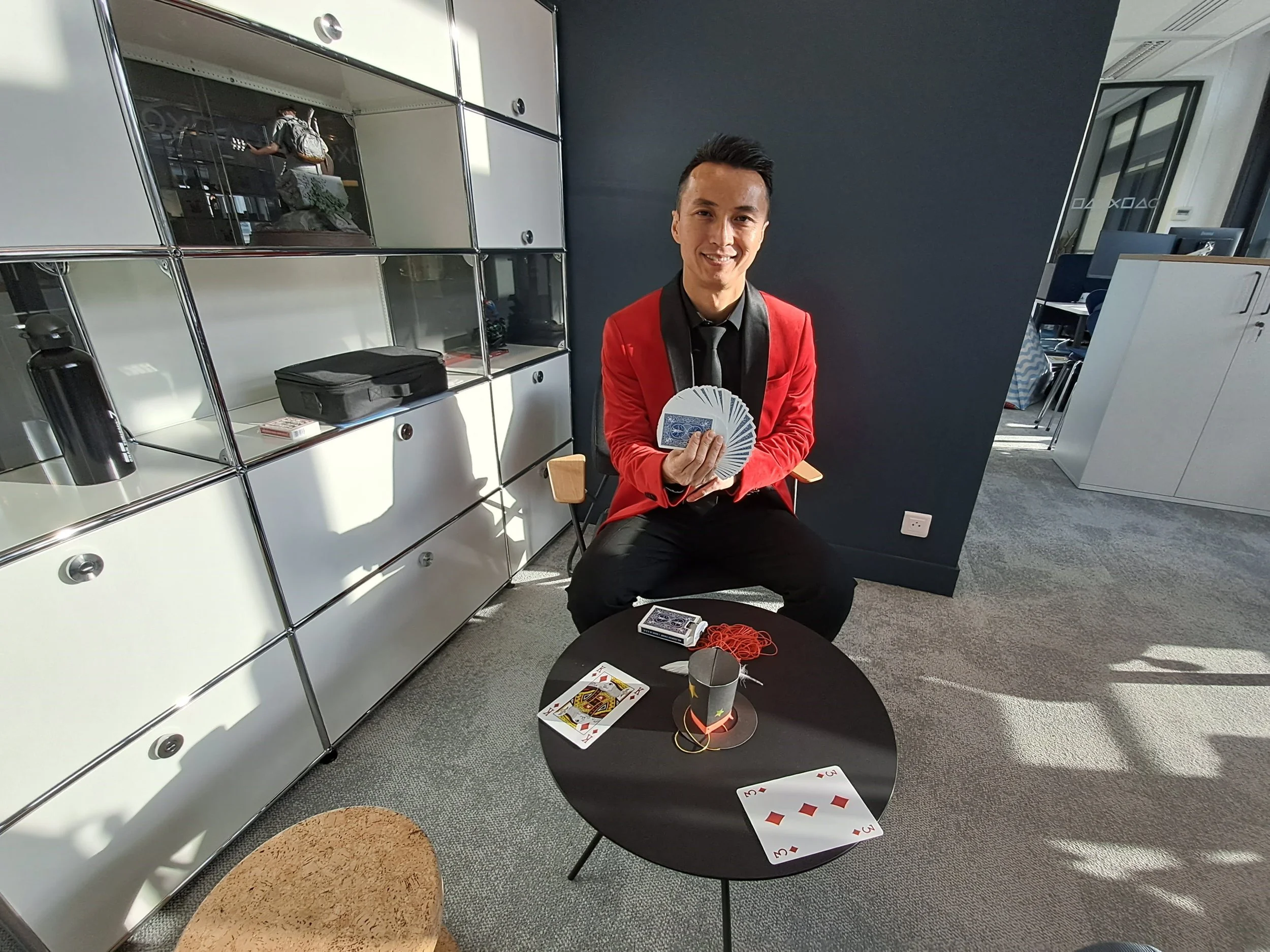 Une personne souriante tenant des cartes à jouer et une balle en fil de coton rouge, assise à une table avec des cartes à jouer, dans un bureau moderne.
