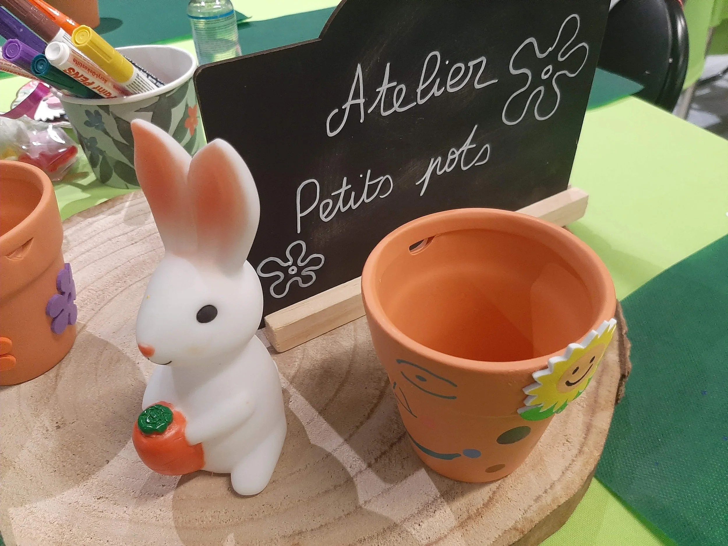 Pot de fleurs décoré avec un motif de soleil, une figurine de lapin en céramique blanche avec une carotte en plastique orange, et un tableau noir avec écriture en français "Atelier Petits pots".
