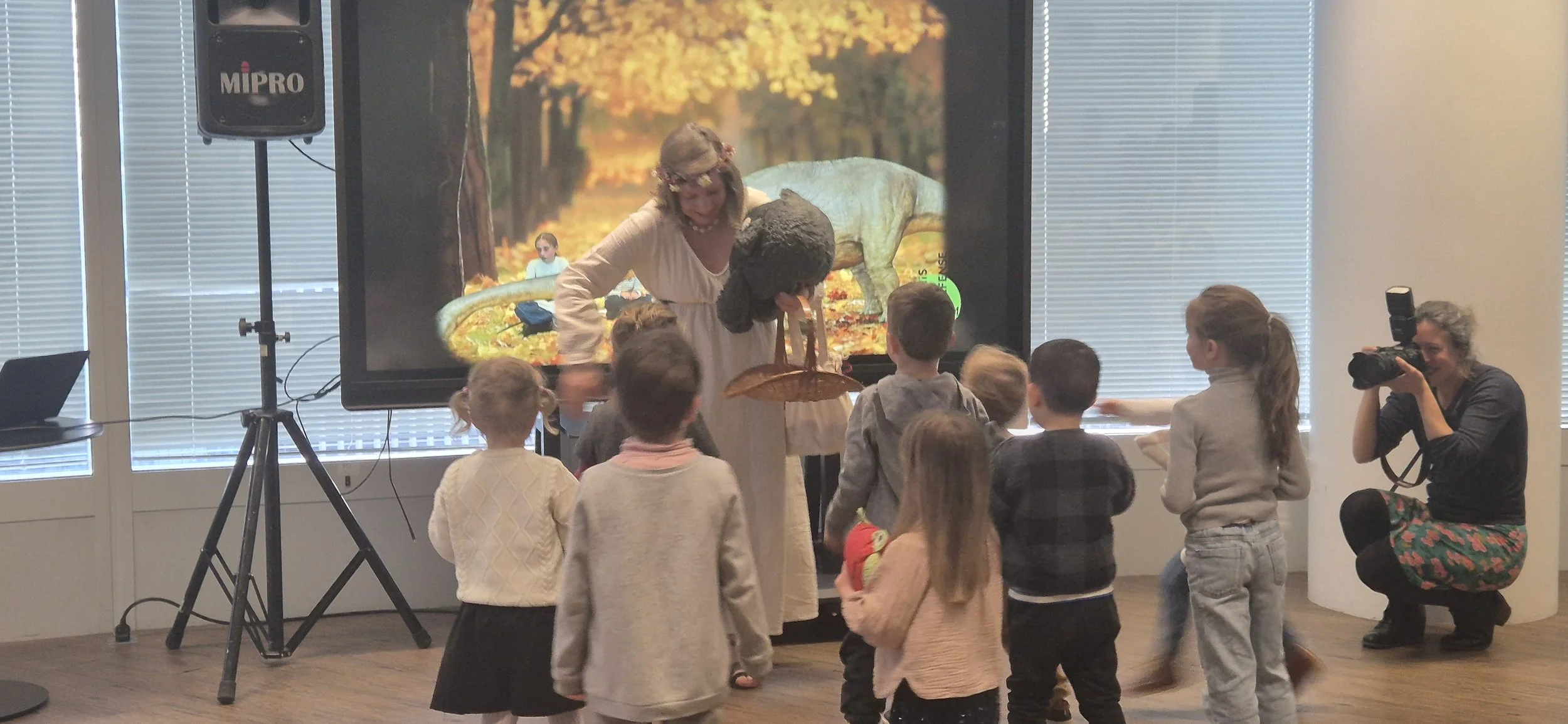 Une femme en costume de conteuse avec des enfants lors d'une activité, en arrière-plan une grande fresque ou écran représentant un paysage de forêt avec un dinosaure.