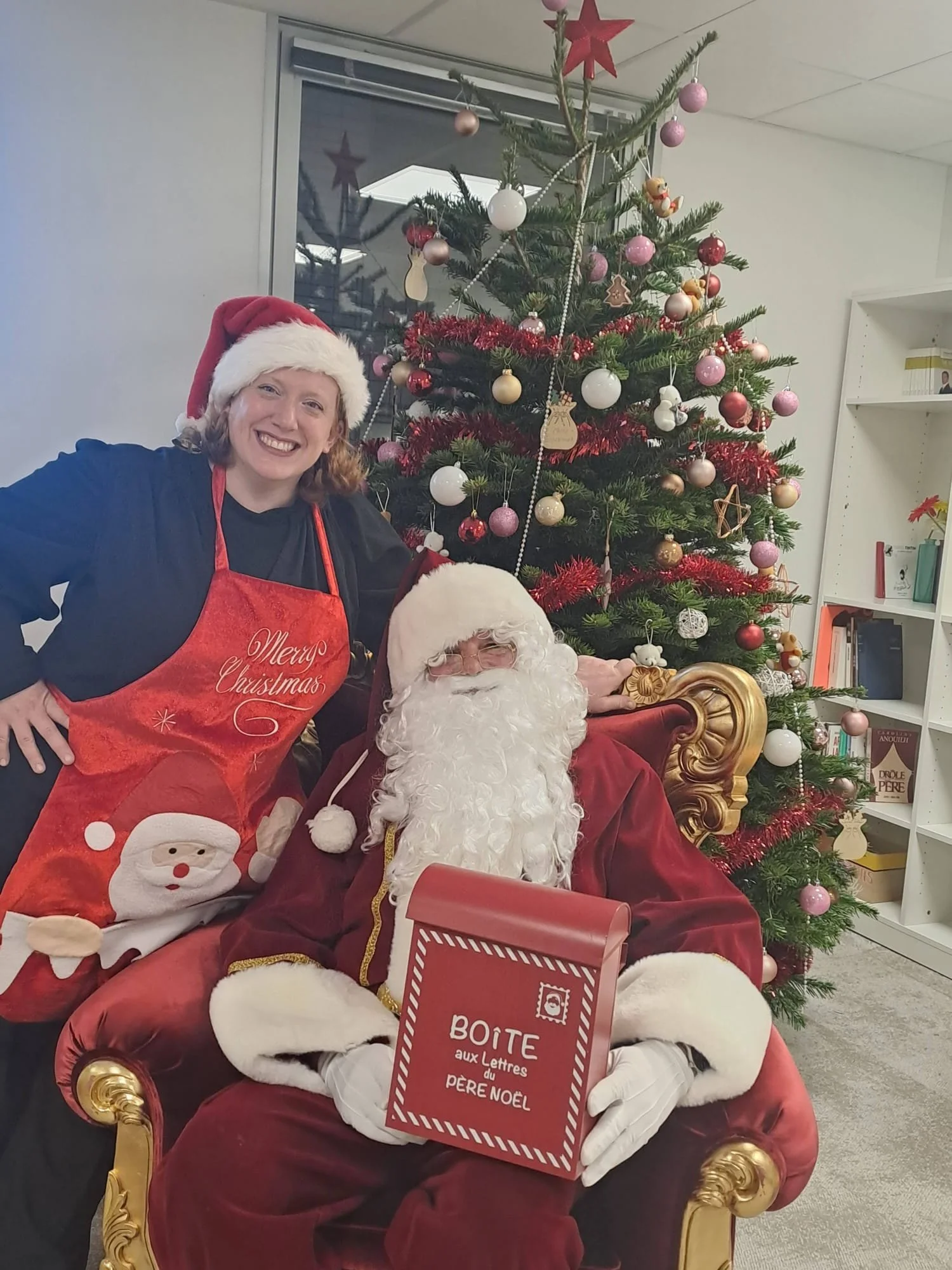 Une femme souriante portant un chapeau de Noël, debout à côté du Père Noël qui est assis sur un fauteuil rouge et doré. Il y a un sapin de Noël décoré avec des boules, des guirlandes et des décorations en forme de biscuits. La scène est dans un bureau ou une bibliothèque, avec des étagères remplies de livres à l'arrière.