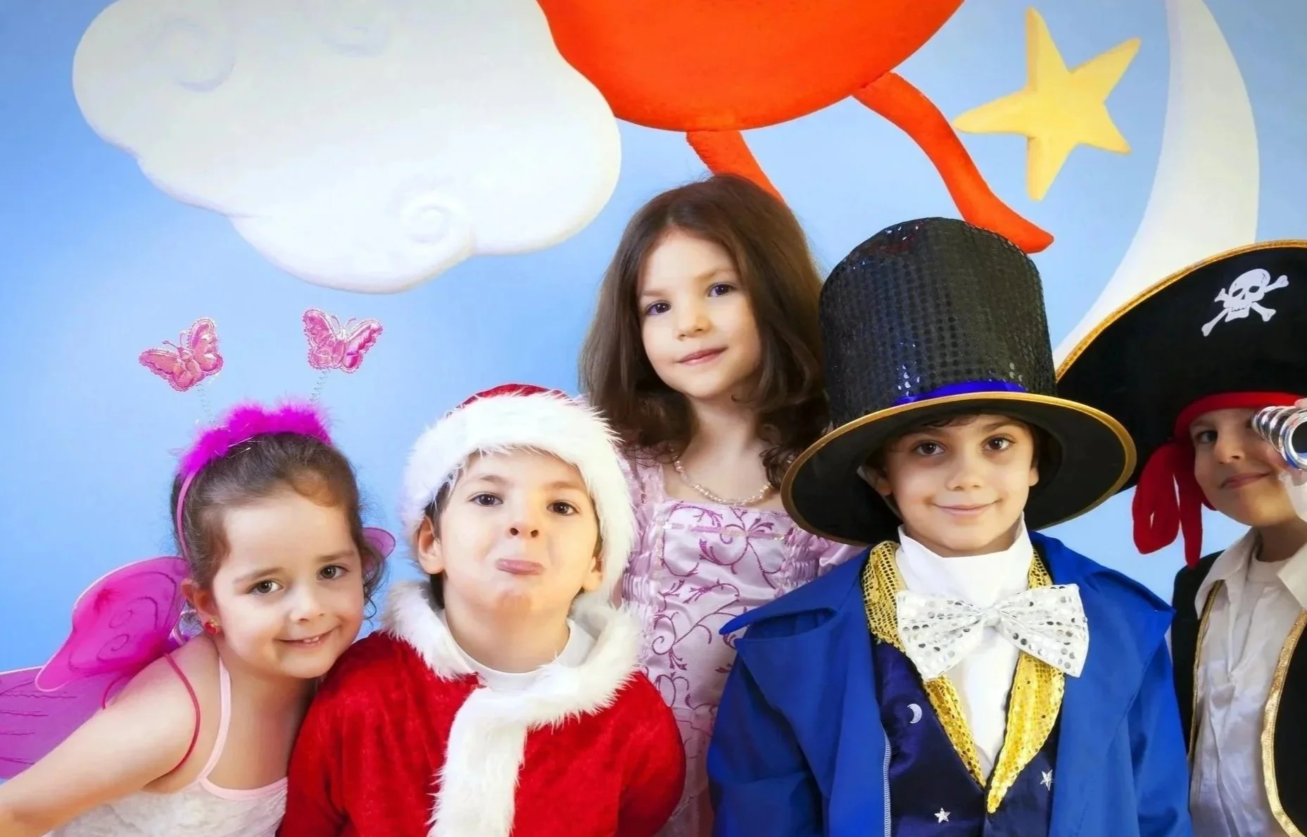 family day  kidsday Groupe d'enfants déguisés pour une fête costumée, avec des costumes de fées, un père Noël, un sorcier et un pirate, devant un décor de ciel avec des nuages, des étoiles et la lune.