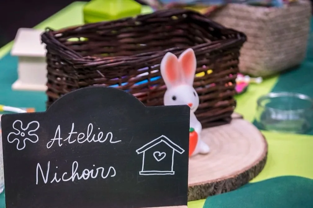 Pot de fleurs décoré avec un motif de soleil, une figurine de lapin en céramique blanche avec une carotte en plastique orange, et un tableau noir avec écriture en français "Atelier Petits pots".