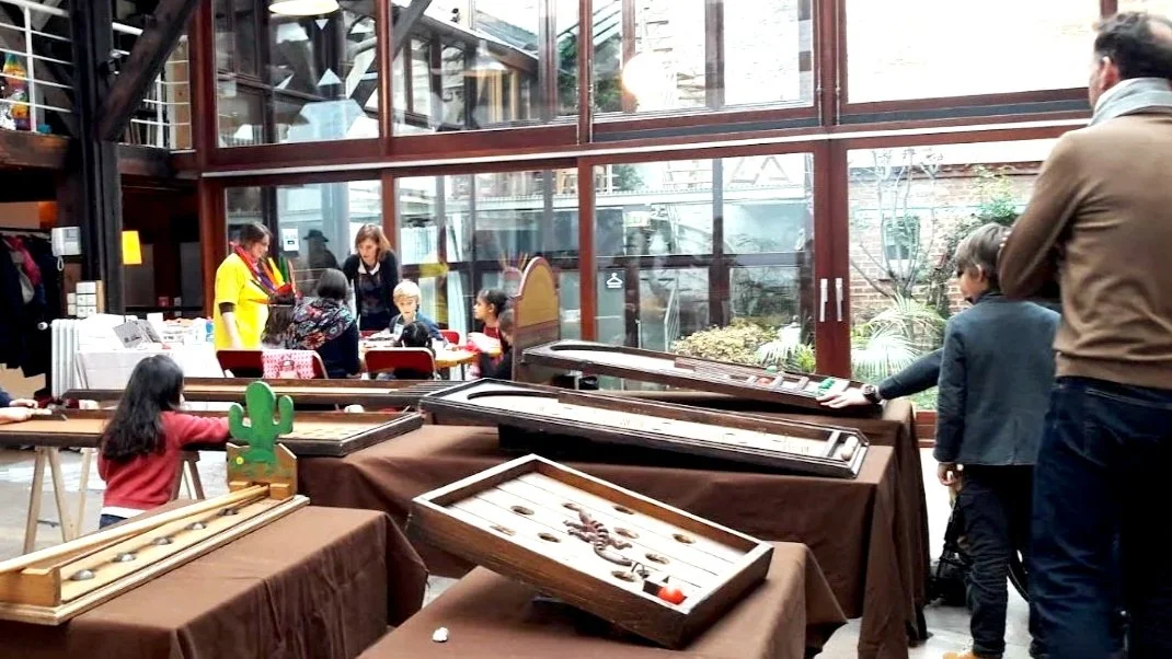 Une salle intérieure avec plusieurs tables recouvertes de nappes marron. Sur certaines tables, il y a des jeux en bois, dont un jeu de morpion et d'autres classiques. Des adultes et des enfants sont présents, certains regardant ou jouant aux jeux, dans un environnement lumineux avec de grandes fenêtres en arrière-plan.