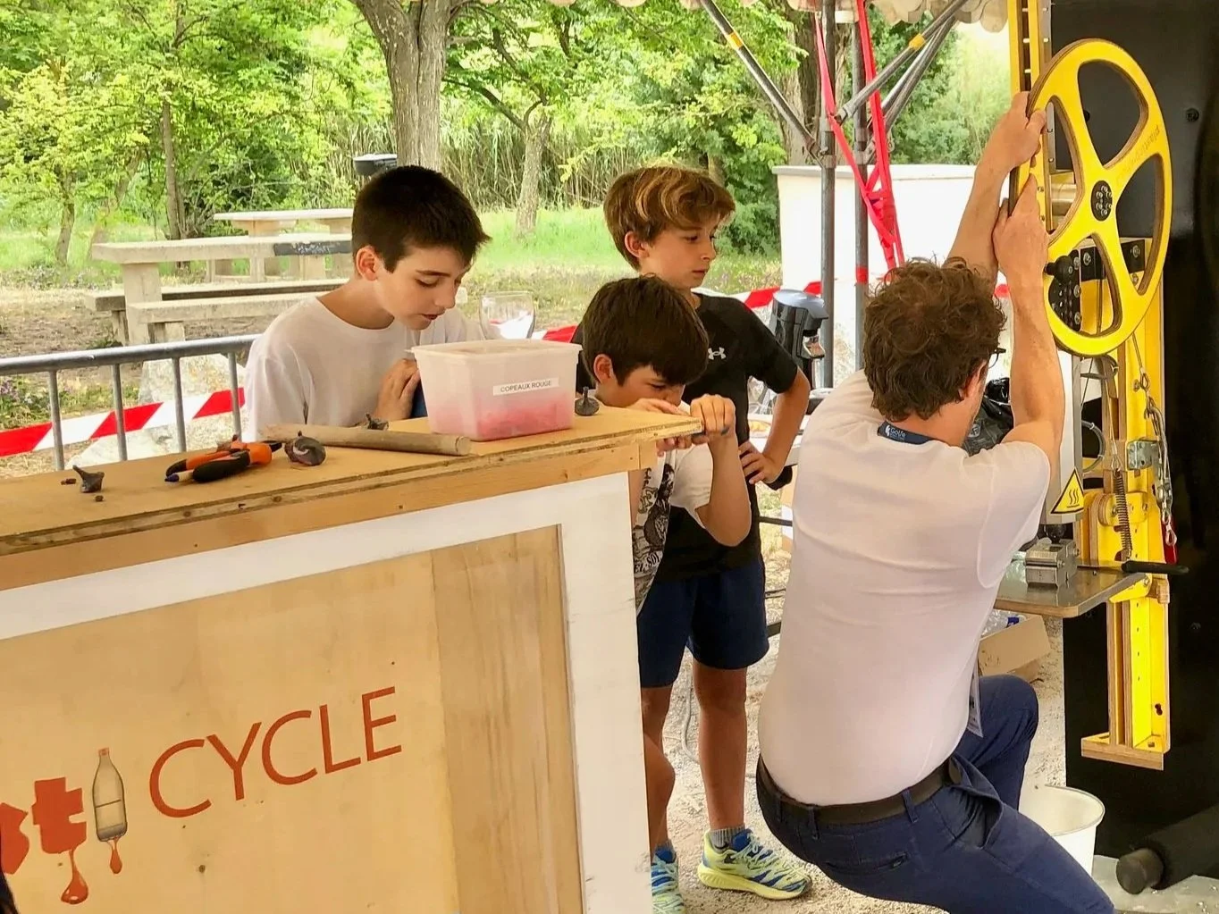 Un homme et trois enfants regardent un mécanisme de machine dans un environnement extérieur sous un abri, avec des arbres et des tables de pique-nique en arrière-plan.