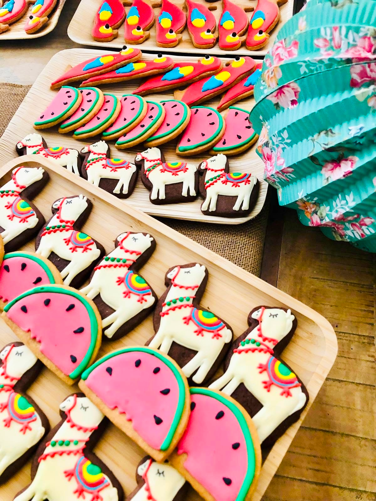 Généreux plateau de biscuits décorés avec des thèmes colorés, comprenant des biscuits en forme de chevaux, de papayes, de flamants roses, et d'autres formes, décorés avec du glaçage coloré et des détails fins, sur des plateaux en bois, à côté d'une boite à cadeaux à motifs floraux.