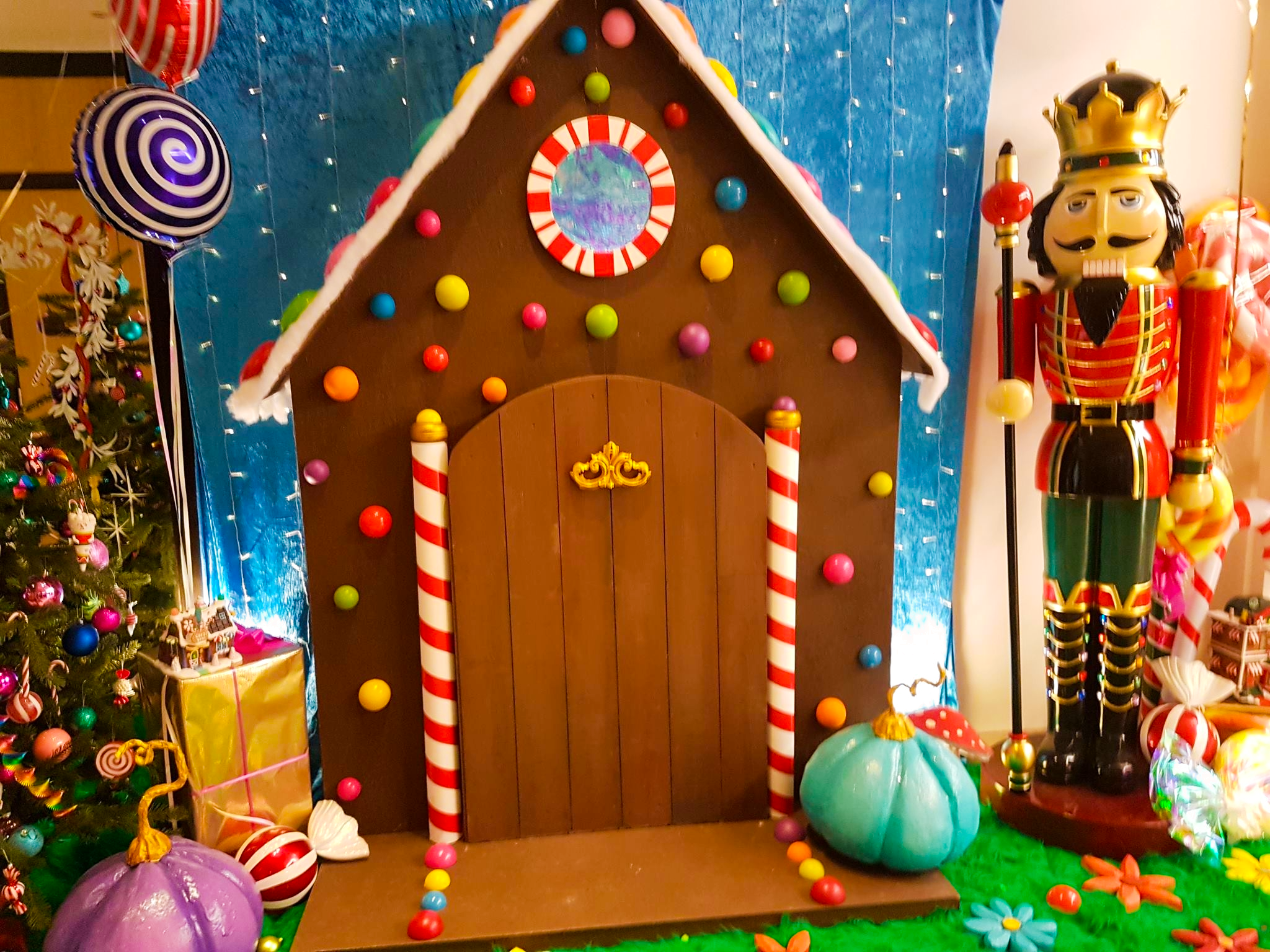 Décoration de Noël avec une petite maison en feutrine, un sapin de Noël miniature, et des paniers de cadeaux et décorations déposés sur une table rouge.