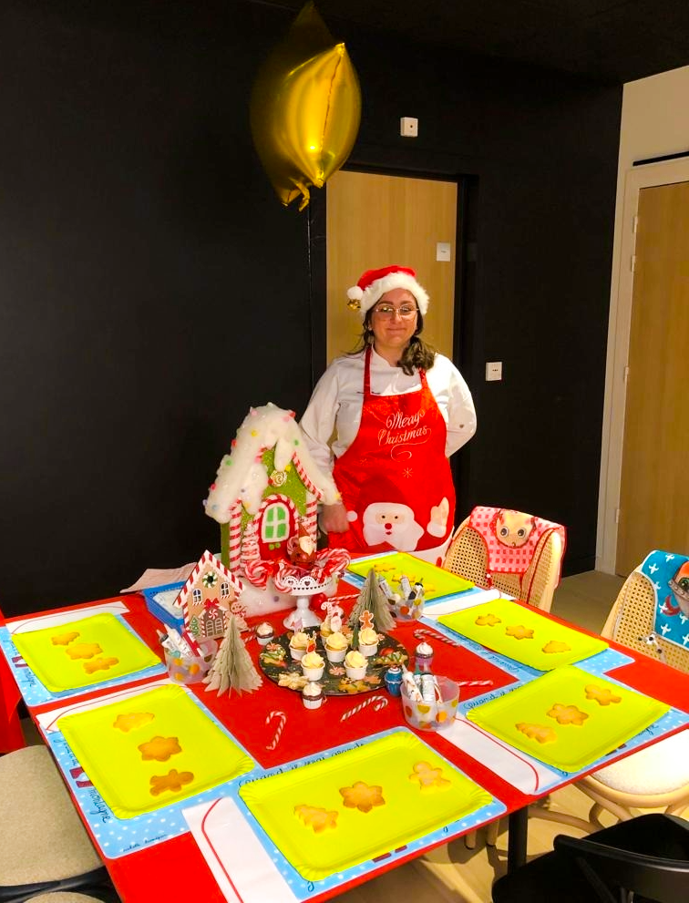 Une femme portant un costume de Noël, avec un chapeau de Père Noël, se tient derrière une table décorée pour Noël avec des cupcakes, des décorations de Noël et un chalet en pain d'épice. Il y a aussi un ballon en forme de ballon de baudruche doré en haut à gauche.