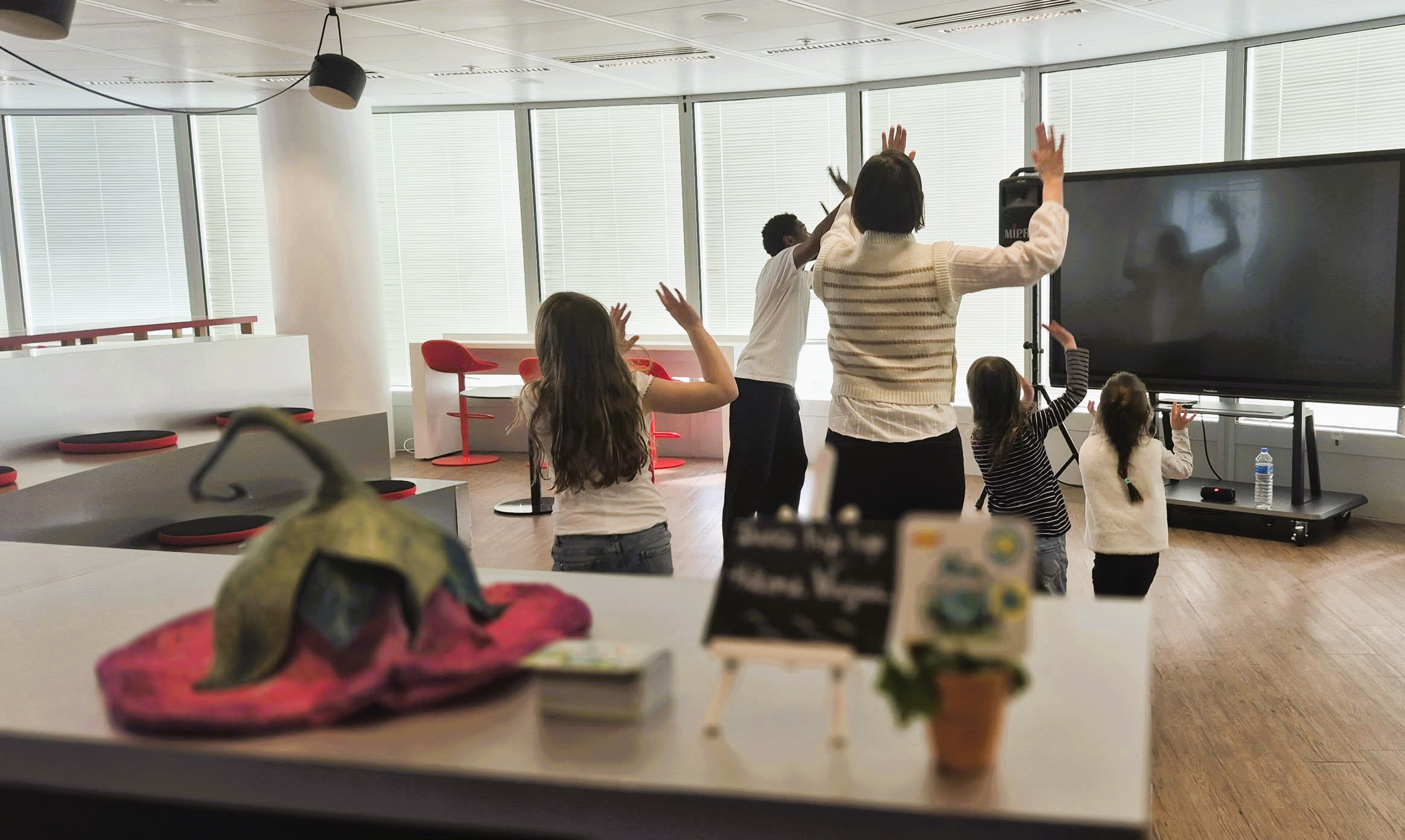 Groupe d'enfants et une adulte en train de faire une activité ou une présentation dans un espace intérieur moderne avec grandes fenêtres et lumière naturelle.