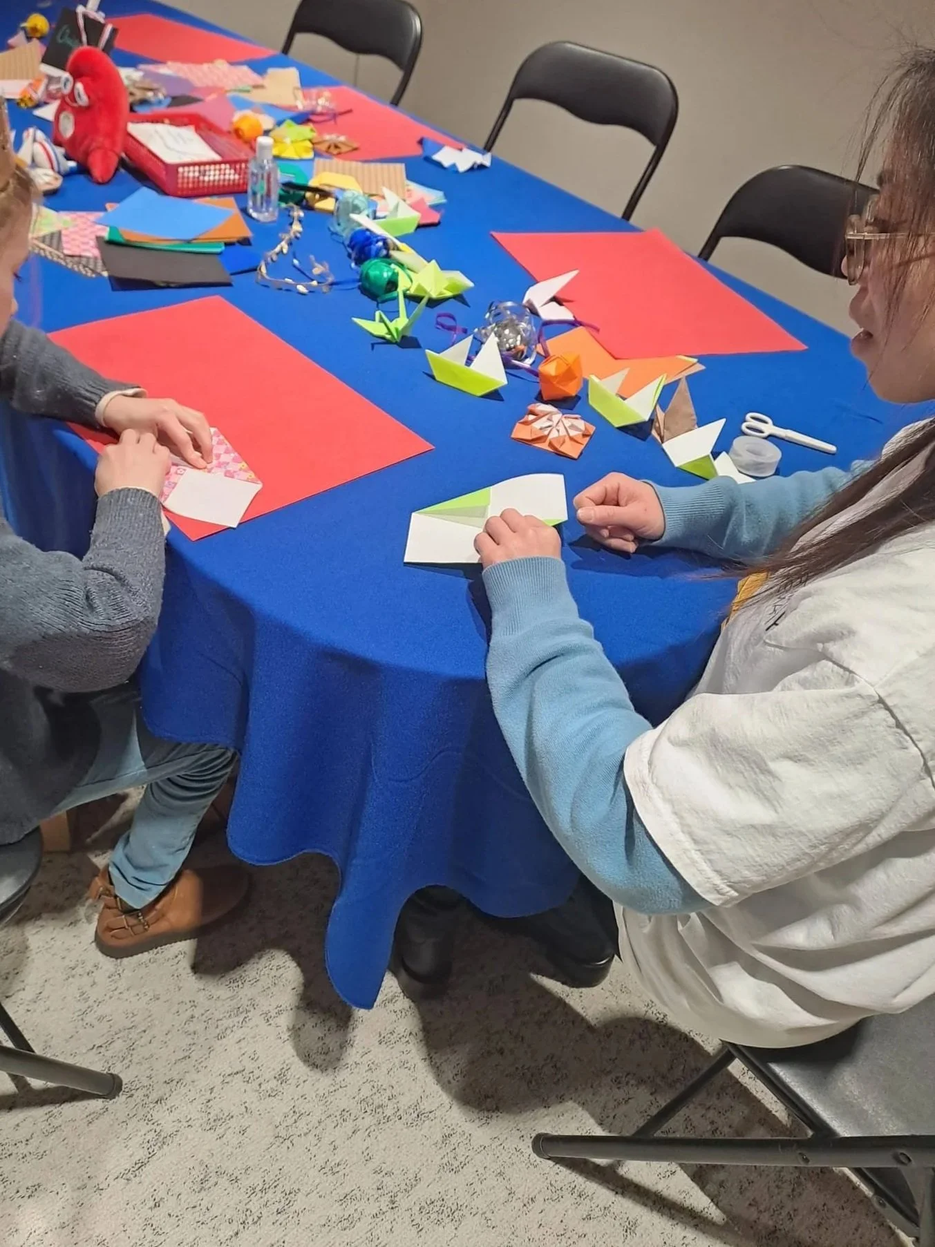 Groupe de personnes assises autour d'une table préparant des pliages en papier, décoration de fête avec des papiers colorés, des origamis, et des objets de décoration.