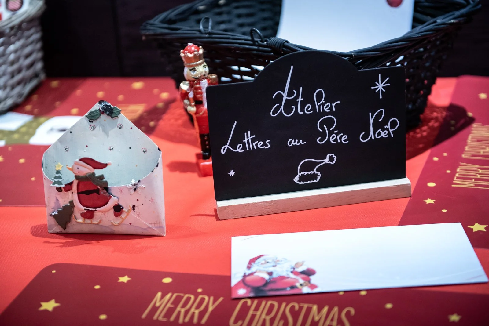 Une table décorée pour Noël avec une petite enveloppe de motif de Noël, une figurine de soldat en bois rouge et blanc, une petite enseigne en ardoise avec l'inscription "Atelier Lettres au Père Noël" et un dessin d'un bonnet de Père Noël, une nappe rouge à motifs d'étoiles dorées et la phrase "MERRY CHRITMAS" écrite en or.