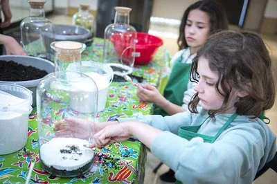 Deux jeunes filles en train d'expérimenter avec des produits de laboratoire sur une table recouverte d'une nappe colorée, avec divers bocaux et flacons contenant des liquides colorés.
