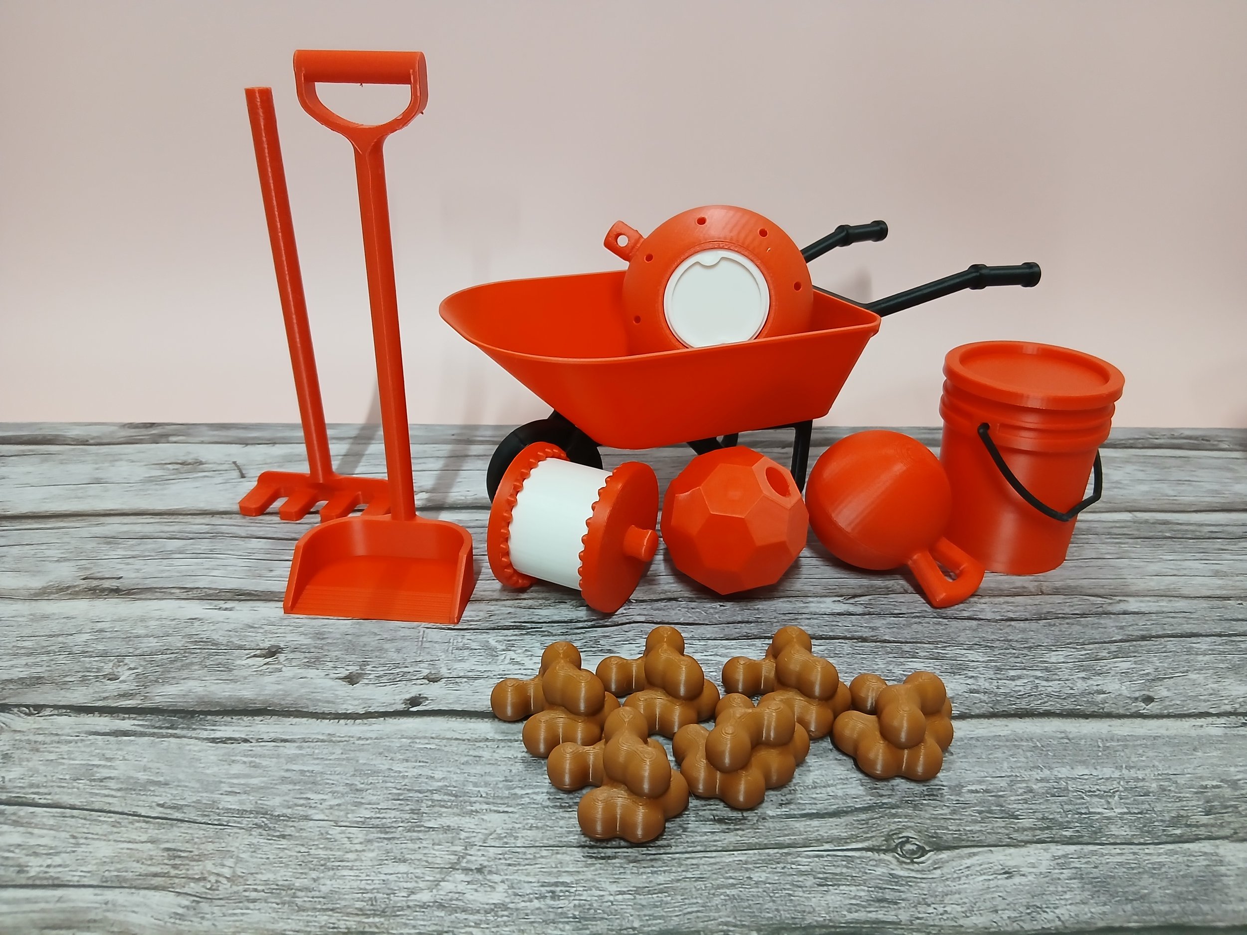 Various orange and black toy gardening tools and accessories including a wheelbarrow, shovel, rake, bucket, and gardening gloves arranged on a wooden surface.
